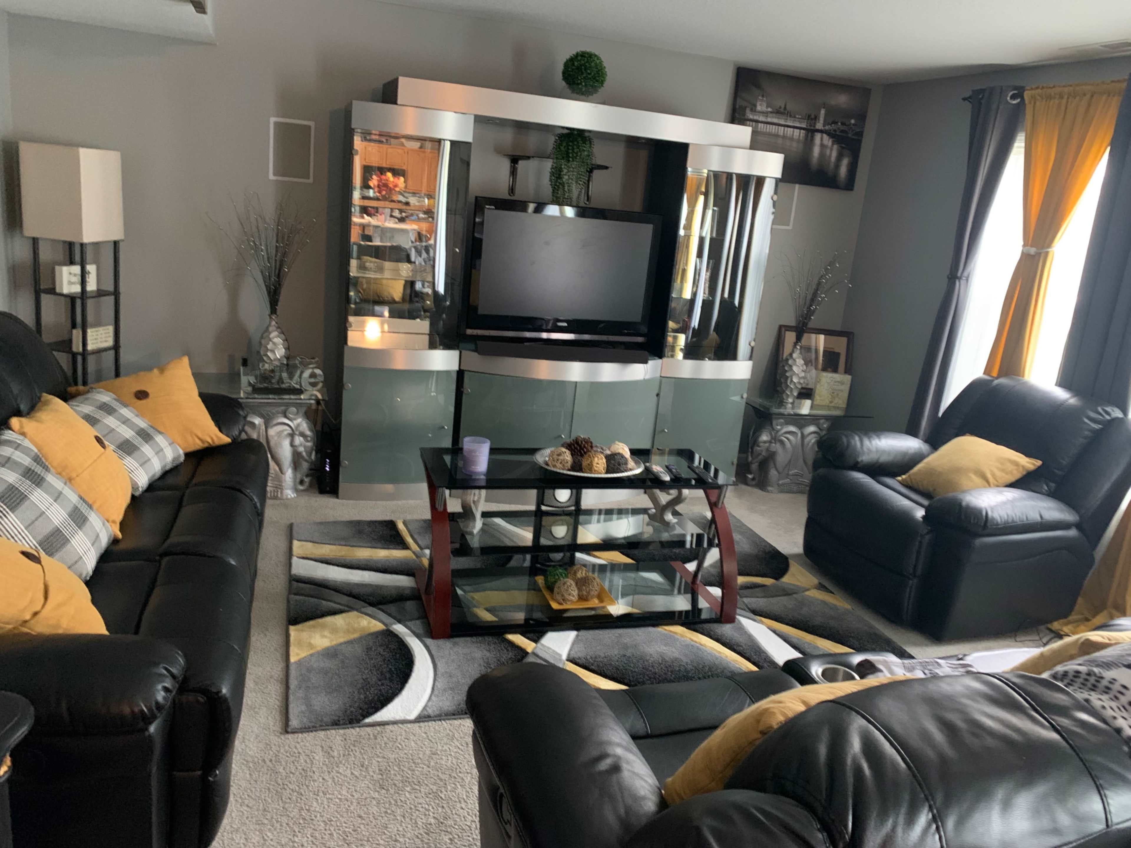 The living room features black leather furniture, a modern glass entertainment center with a television, and decorative accents on a patterned area rug.
