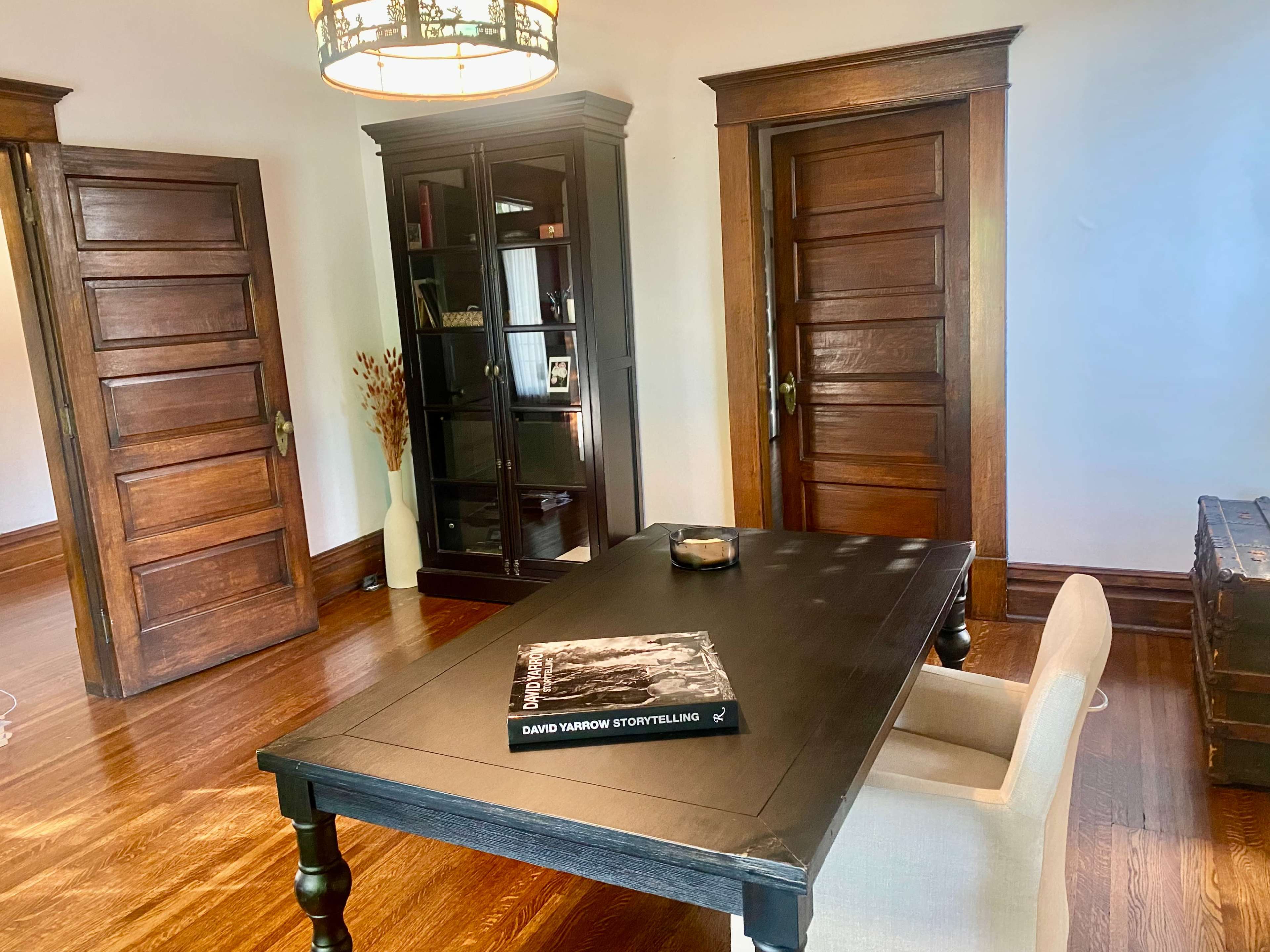 The image shows a room with a black table, a light-colored chair, and two wooden doors, along with a glass-front cabinet and a book on the table.