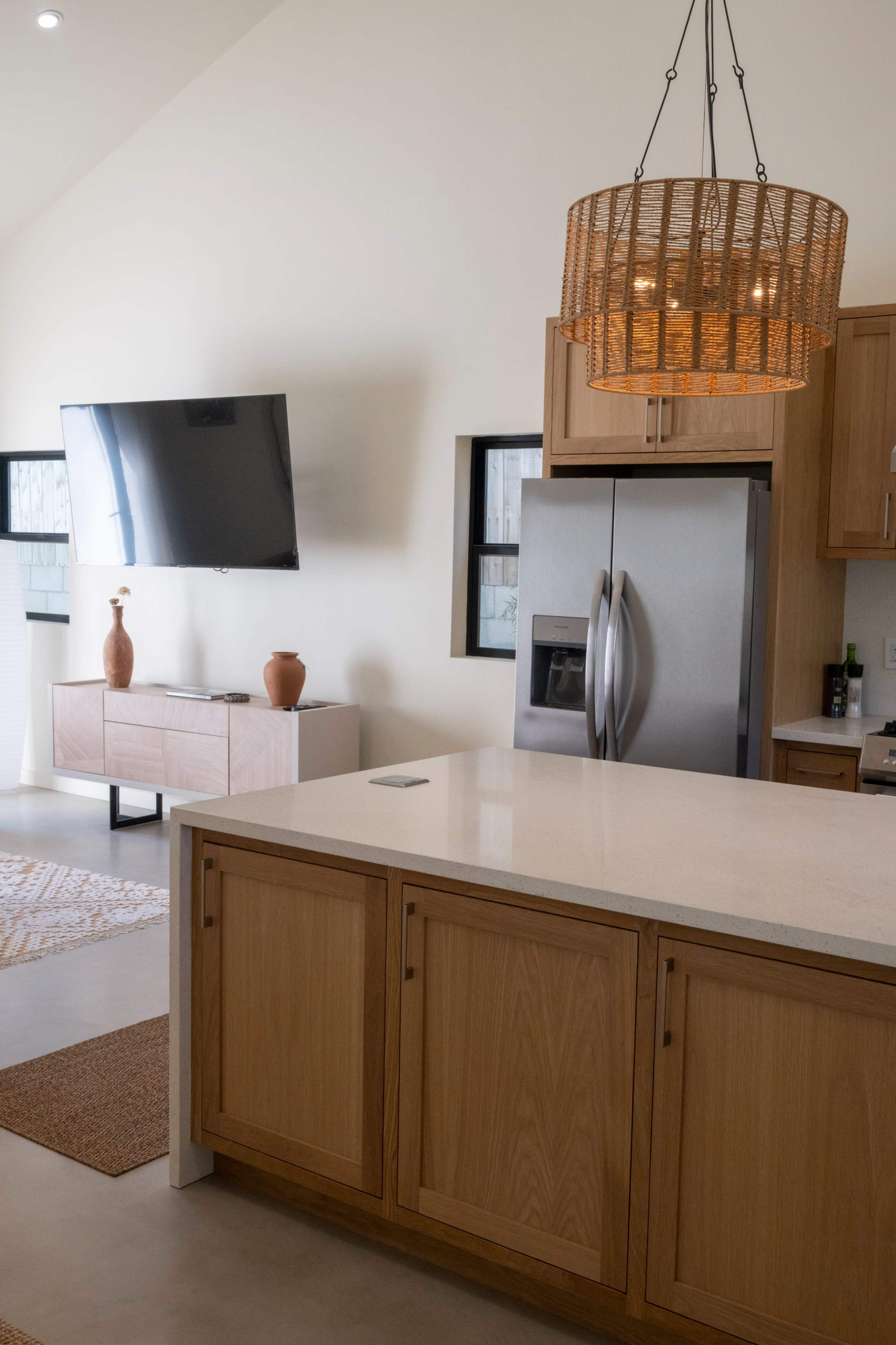 The image depicts a modern kitchen featuring wooden cabinetry, a stainless steel refrigerator, a large flat-screen TV mounted on the wall, and a woven chandelier hanging from the ceiling.