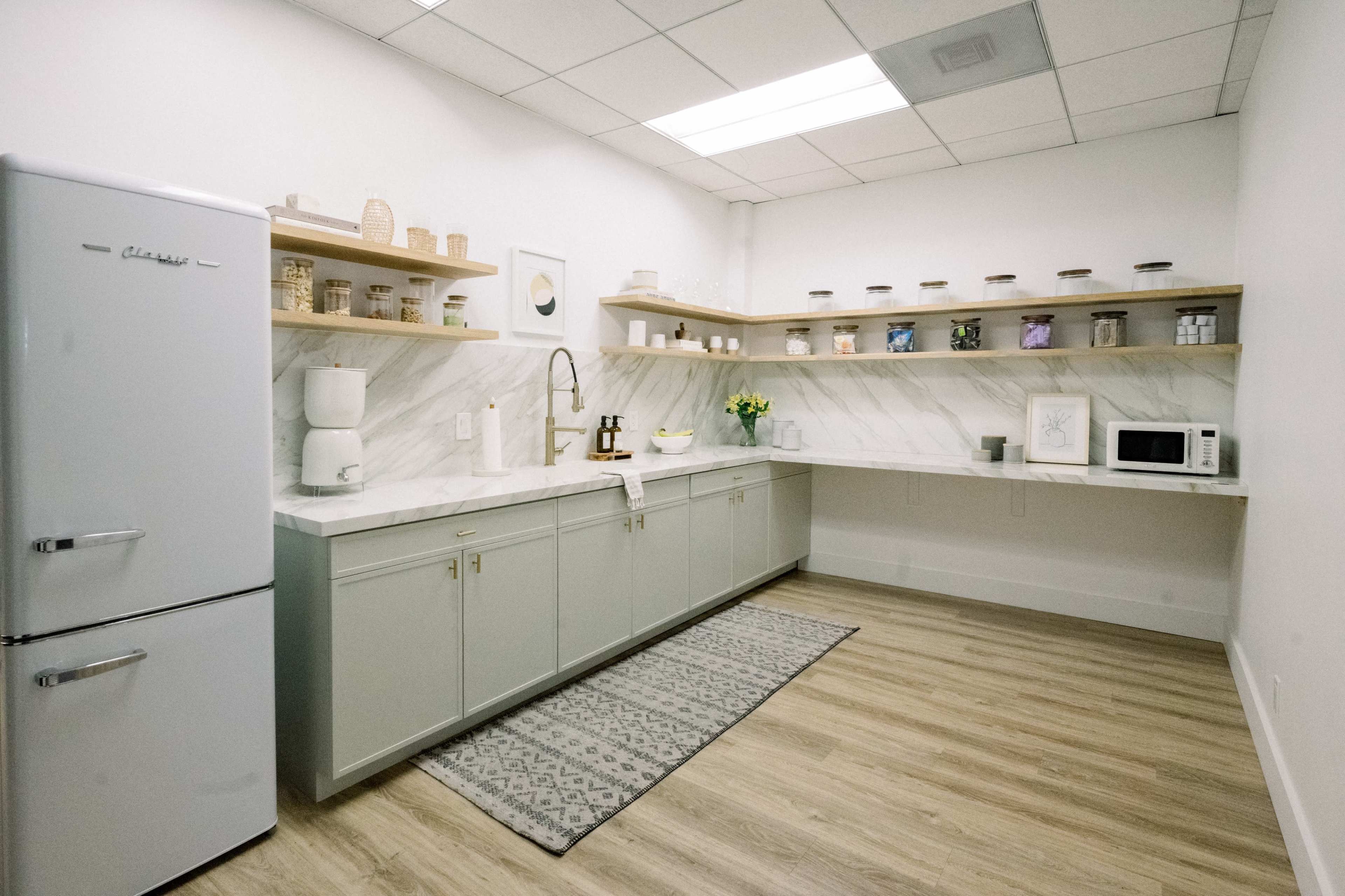 The image shows a modern kitchenette with light gray cabinets, a marble countertop, and shelves displaying jars, along with a refrigerator and microwave.