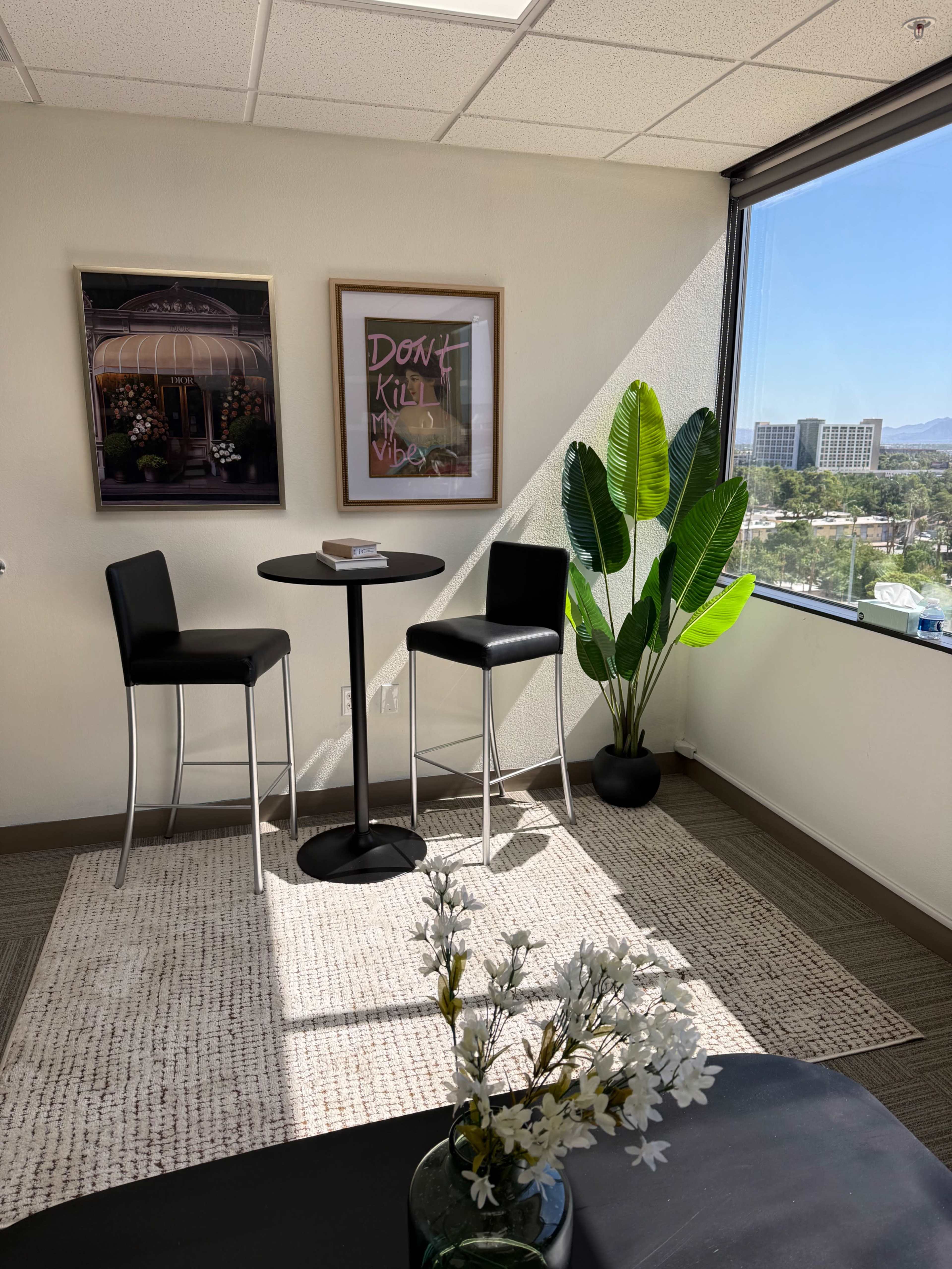 The image shows a bright office corner with two black high stools, a round table, a potted plant, and a decorative rug, all positioned near a large window.