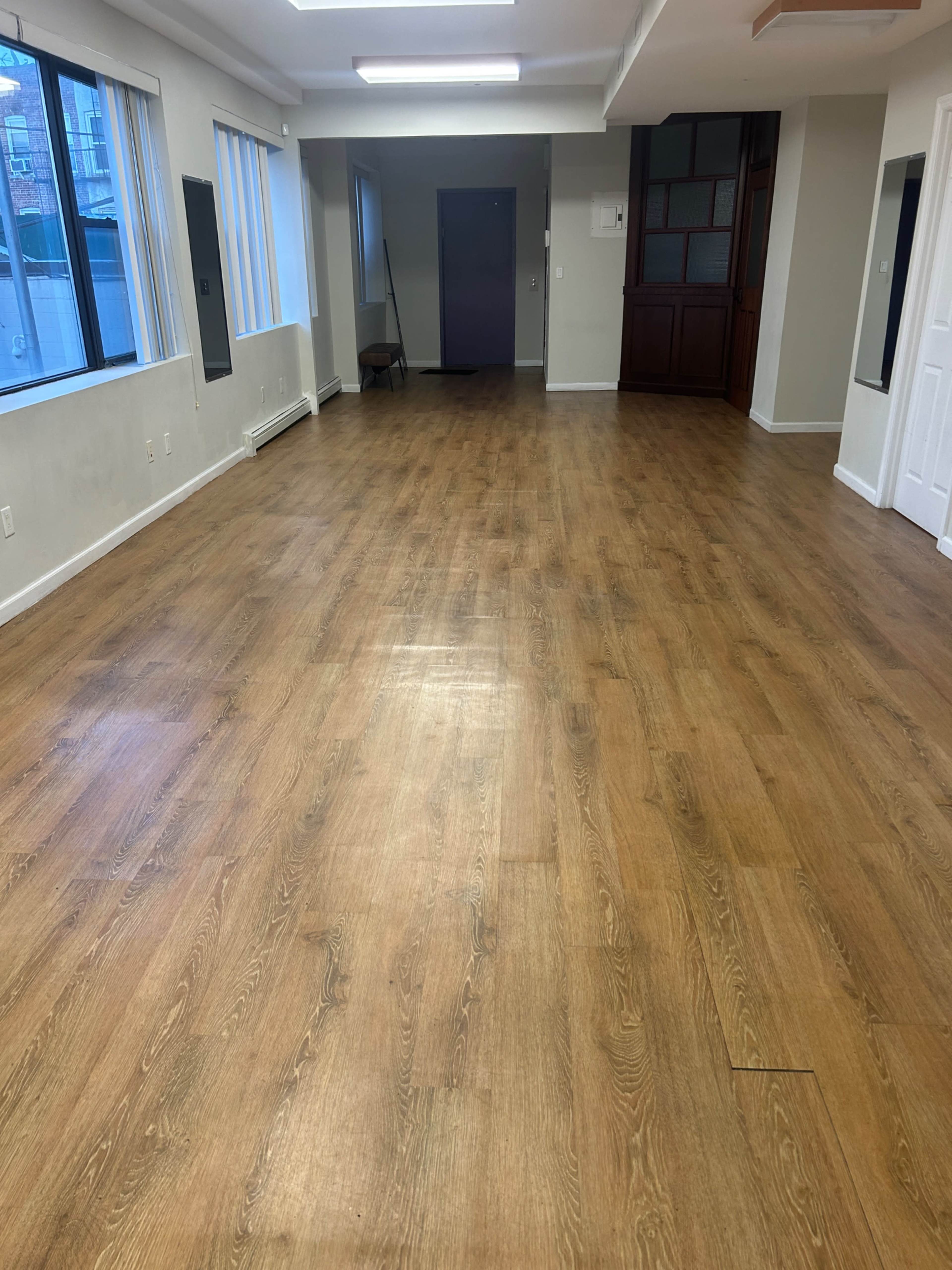 The image shows a spacious, empty room with wooden flooring and large windows lining one wall.