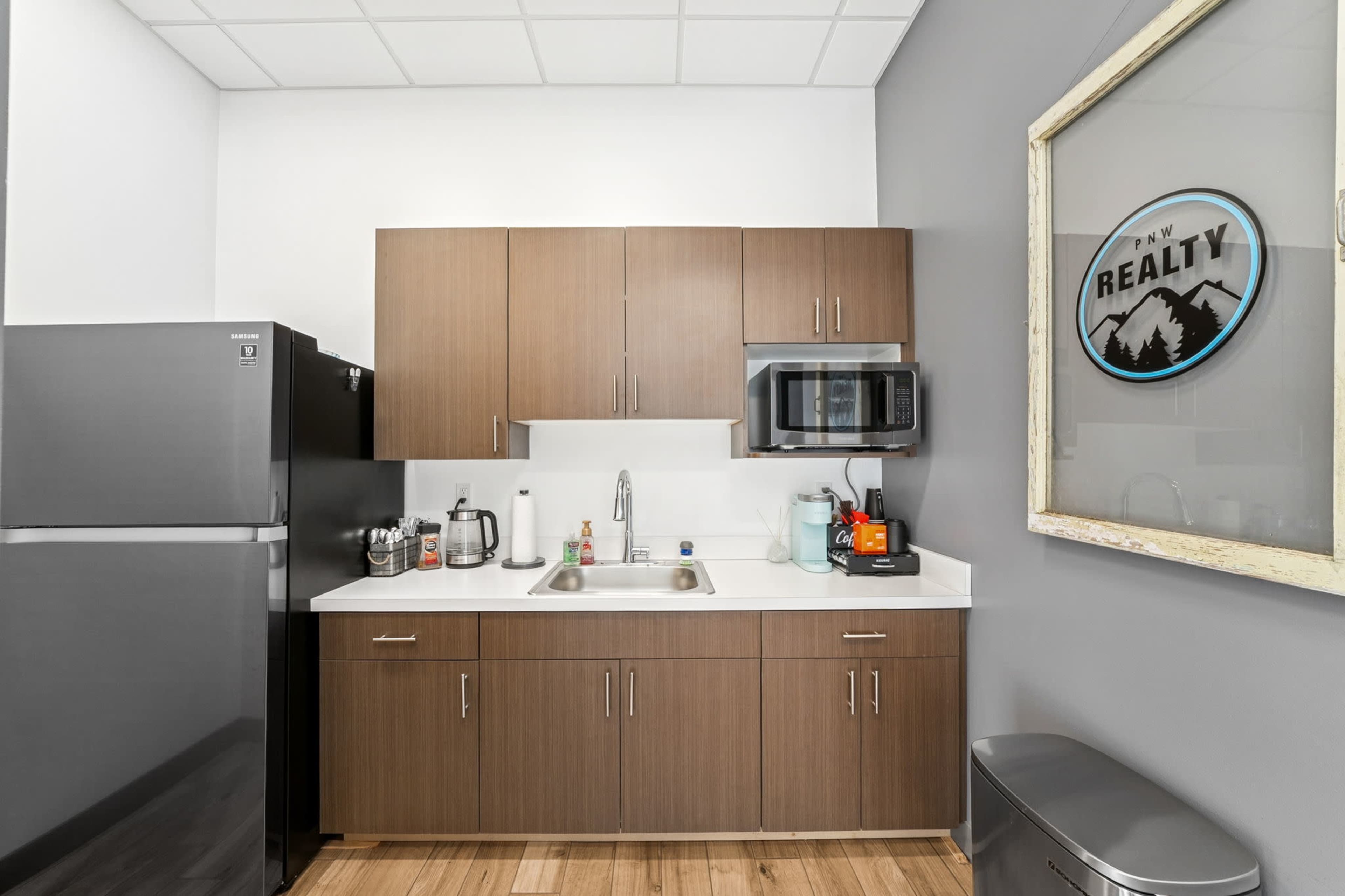 The image shows a modern kitchenette with wooden cabinets, a stainless steel refrigerator, a microwave, and a sink, along with various kitchen appliances and utensils.
