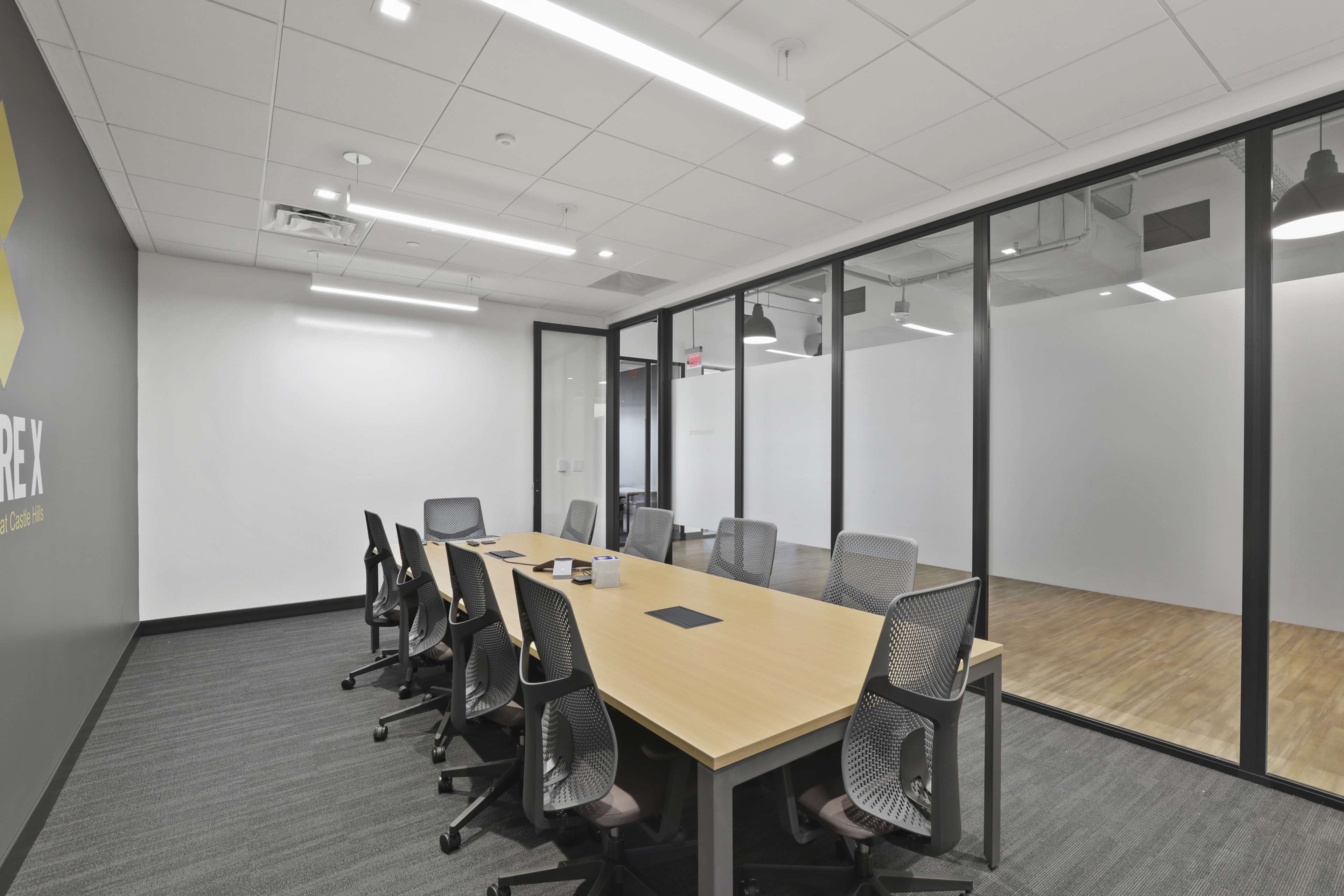 A modern conference room features a large wooden table surrounded by ergonomic chairs, with glass walls separating it from an adjacent space.