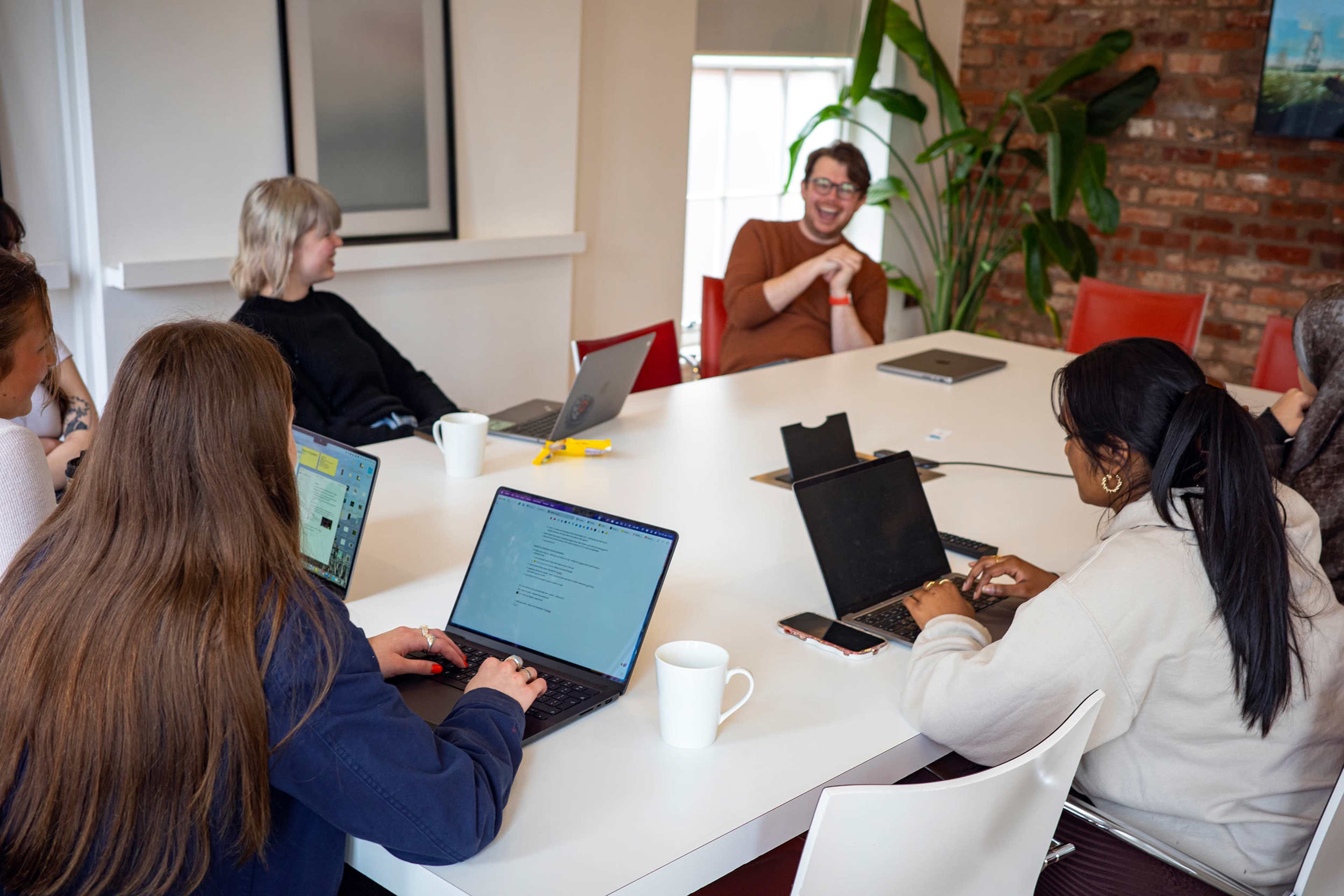 A group of eight people are seated around a table in a well-lit room, engaging in discussion while using laptops.
