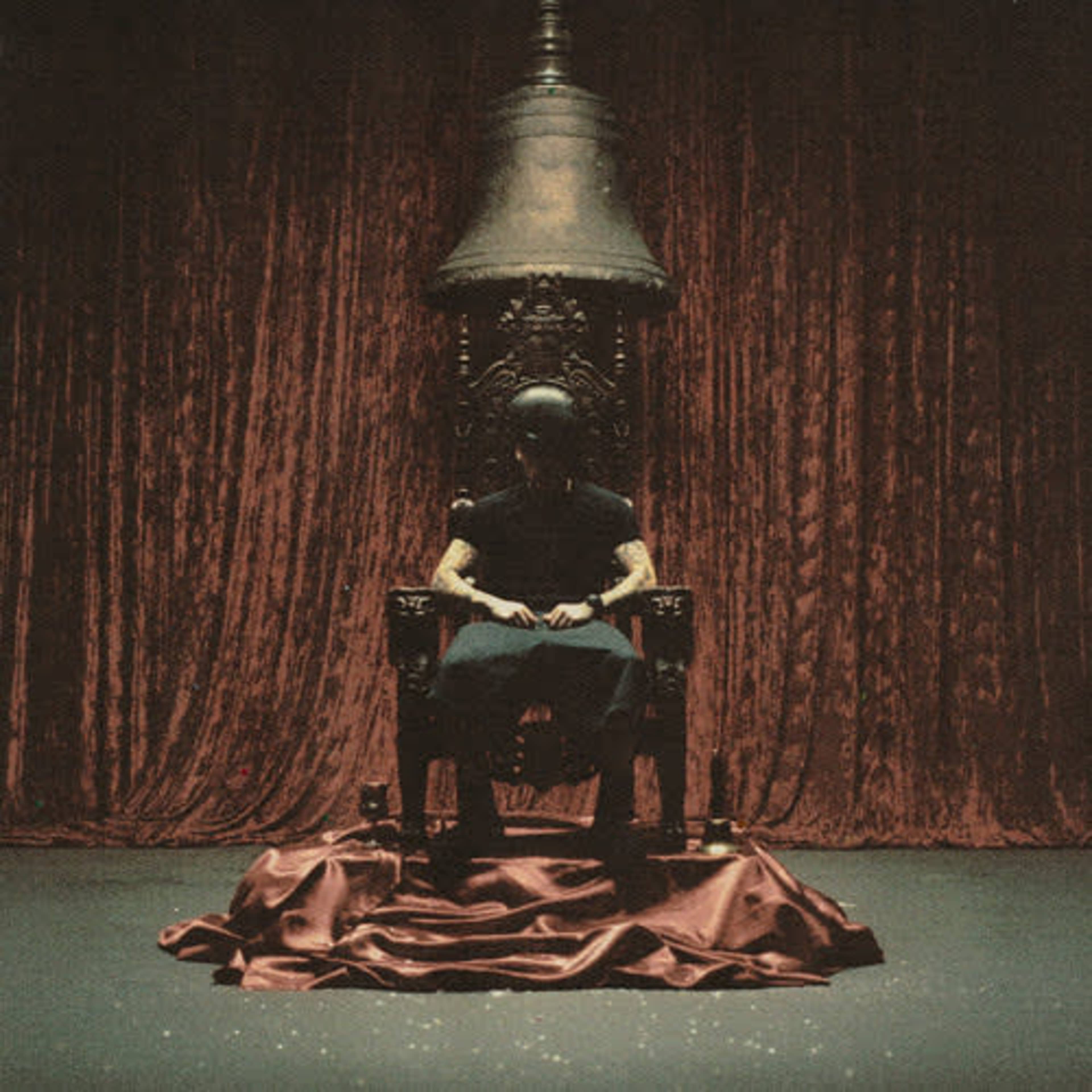 A figure sits on an ornate throne draped with red fabric in front of a heavy curtain.