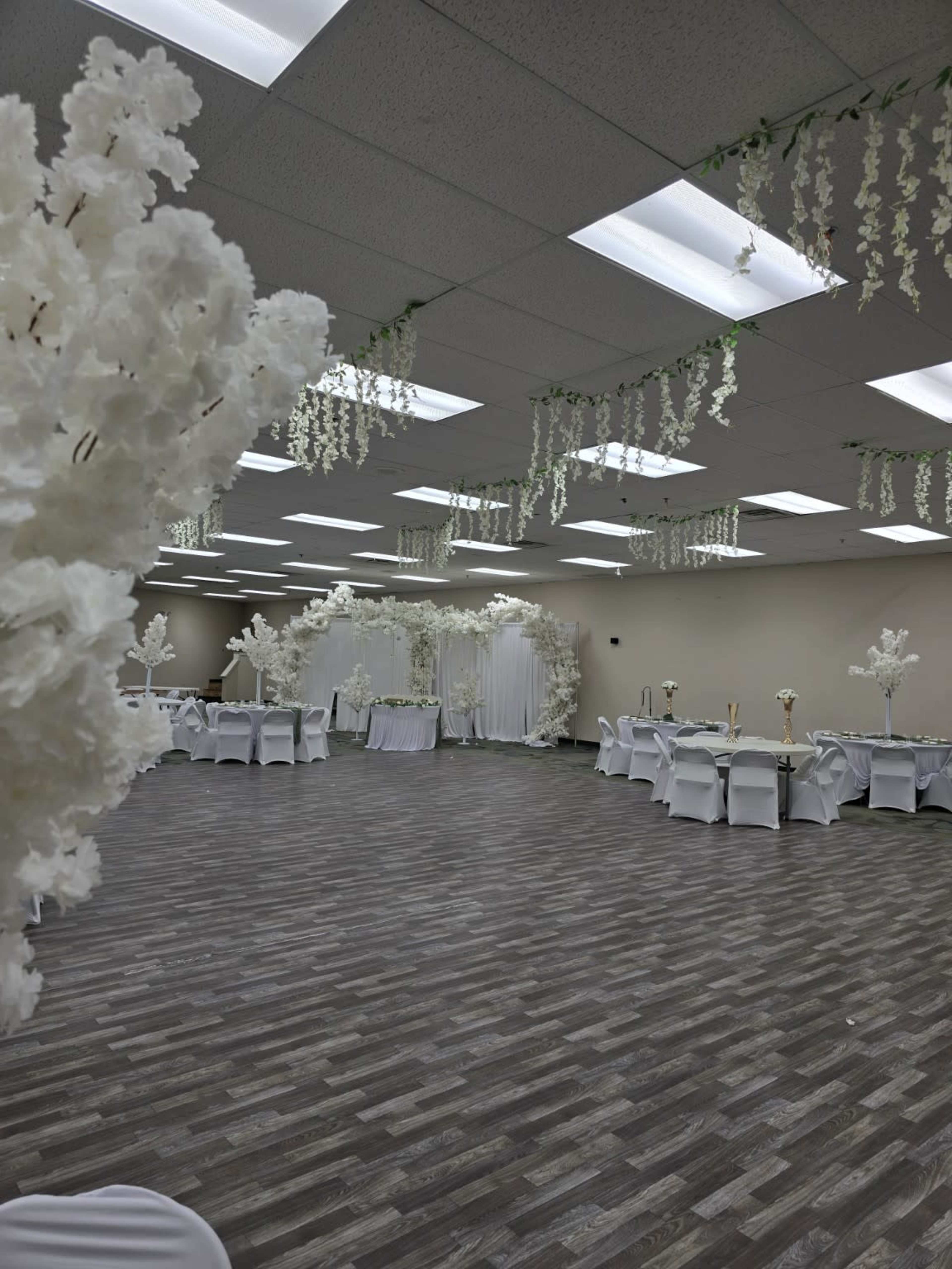 The image shows a spacious event hall decorated with white floral arrangements and draped greenery hanging from the ceiling, set with tables covered in white tablecloths.