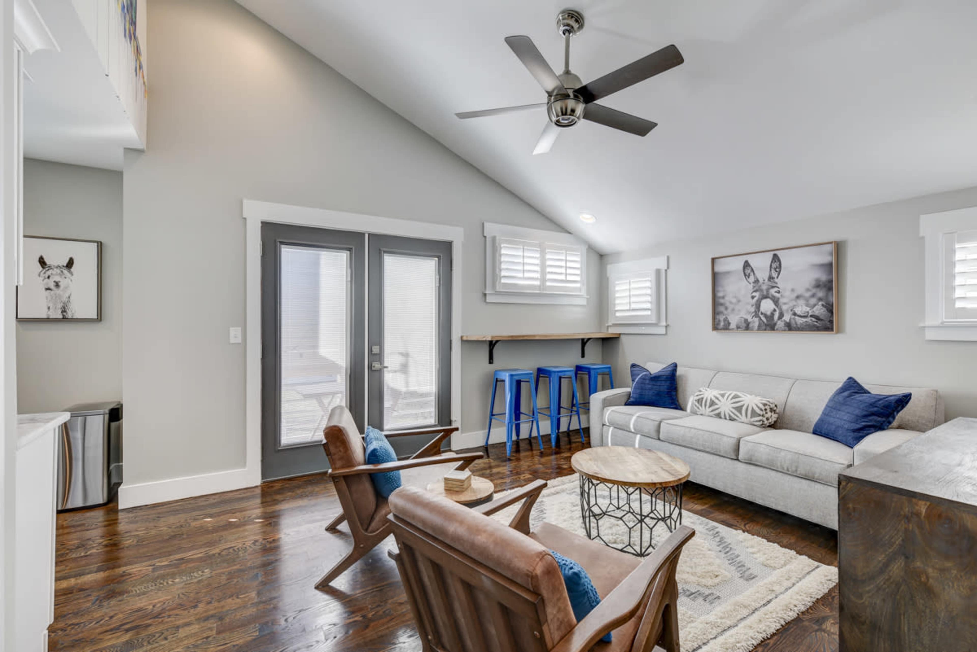 A living room with a gray sofa, wooden chairs, a round coffee table, and a bar area with blue stools, all set against light-colored walls and hardwood flooring.