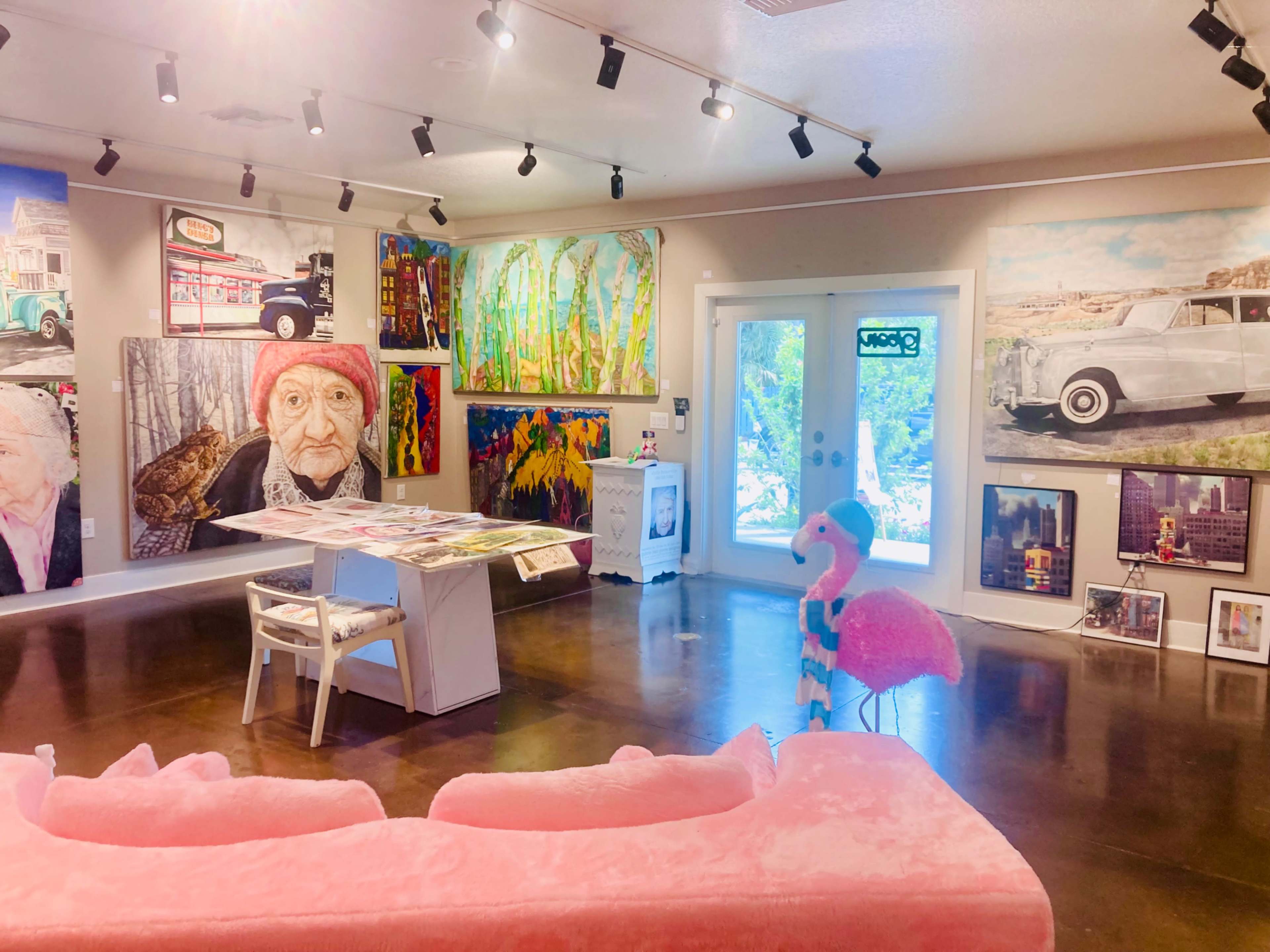 The image shows an art gallery featuring various colorful paintings on the walls, a white table with art materials in the center, and a pink flamingo sculpture near a plush pink sofa.