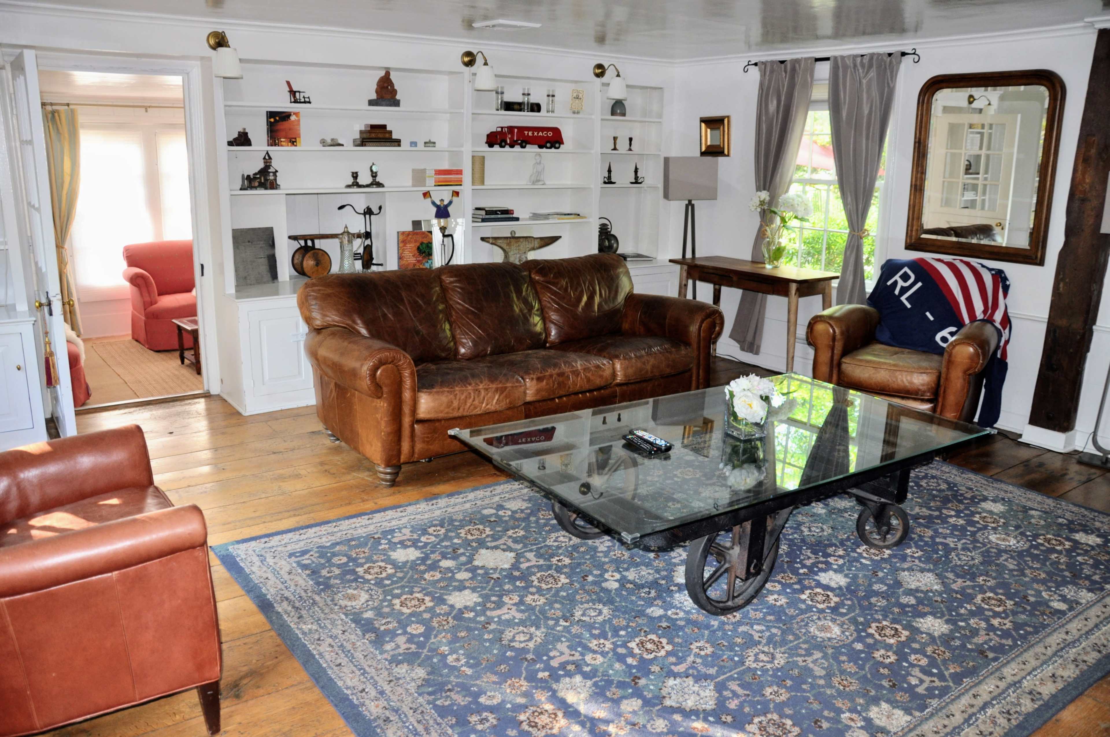 The image shows a living room featuring a leather sofa, a glass coffee table on a patterned rug, and bookshelves filled with various decorative items.