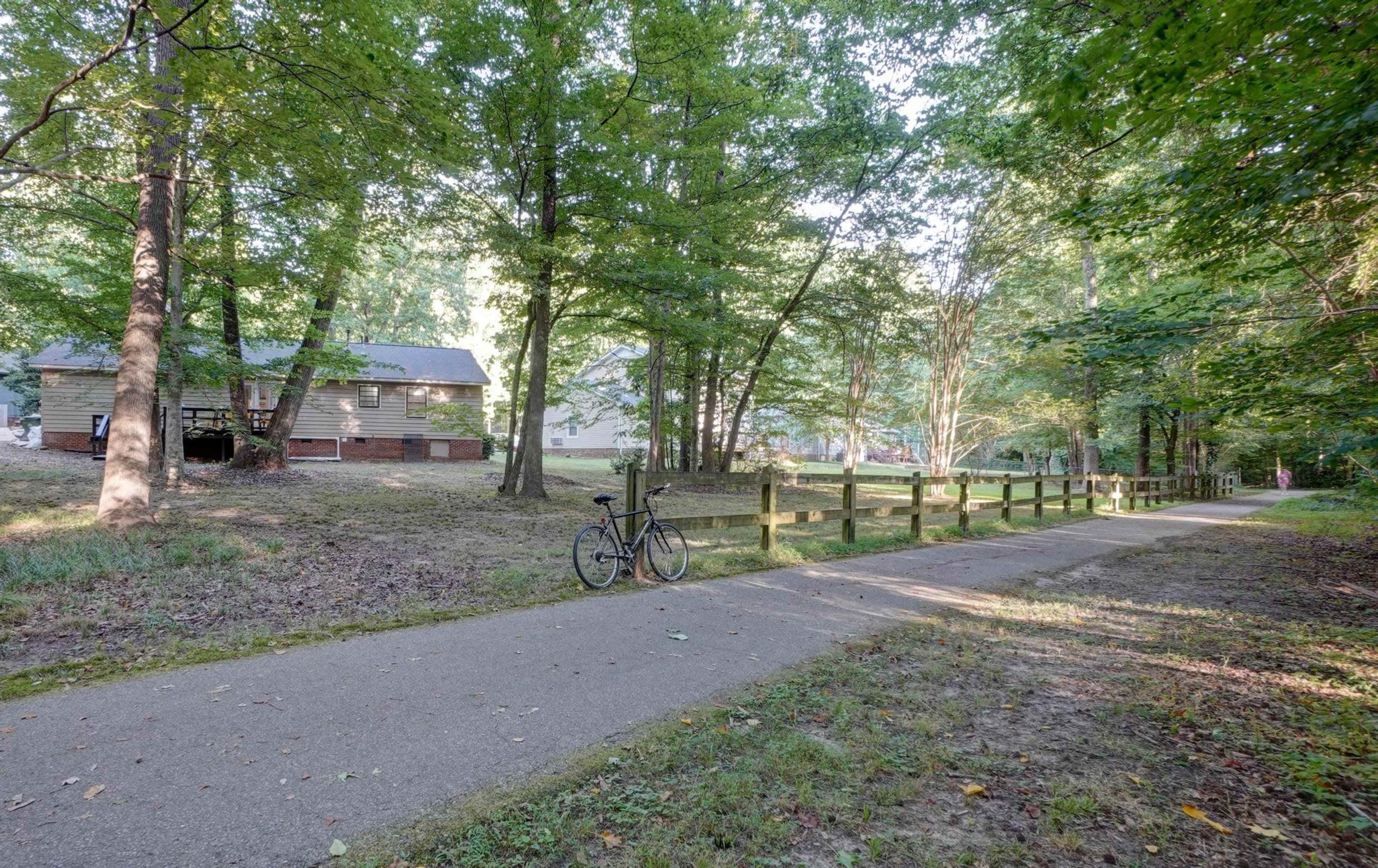 A paved path runs beside a wooden fence in a wooded area, with a bicycle parked nearby and a house visible in the background.