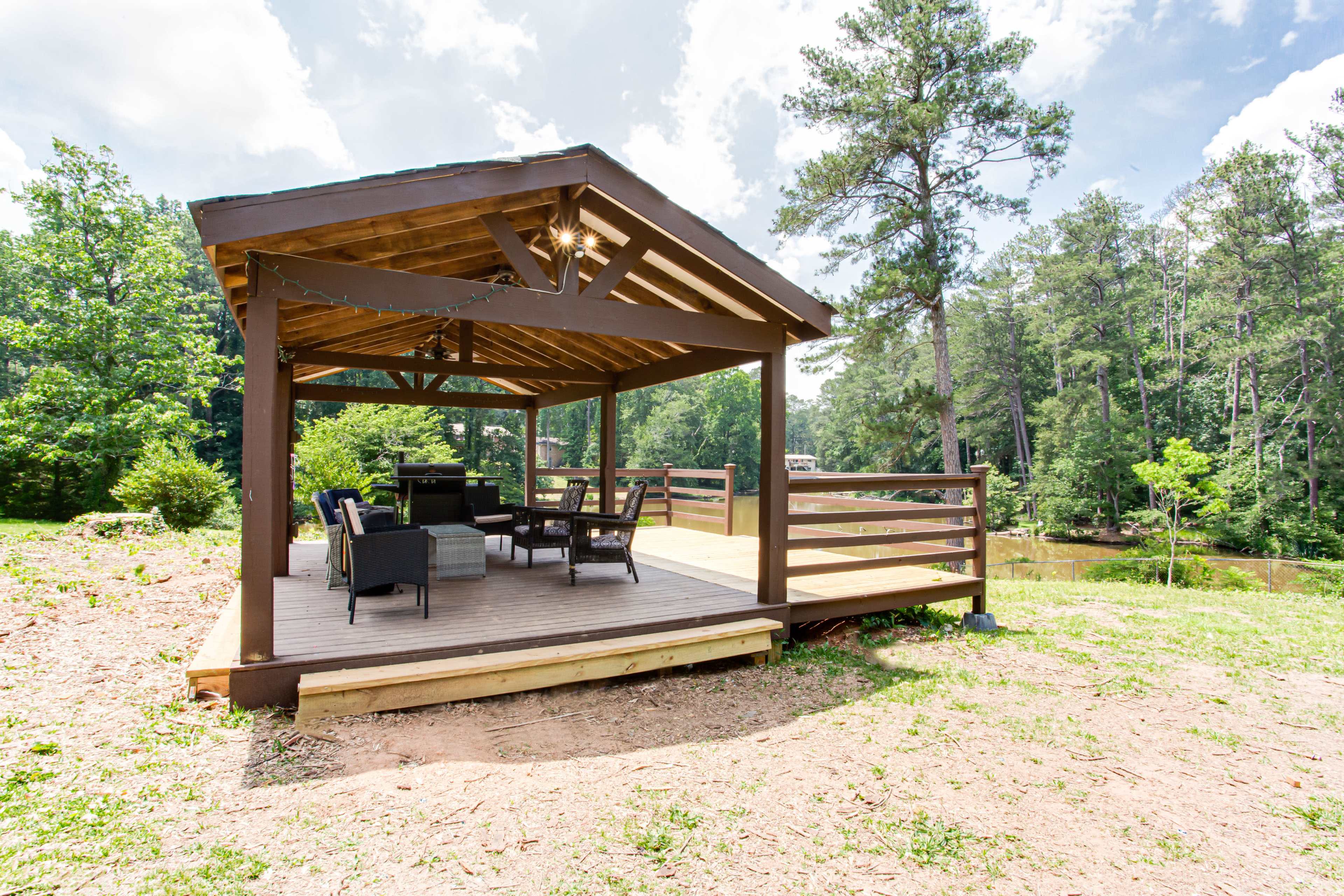 A covered patio with seating is situated next to a small pond, surrounded by tall trees and greenery.
