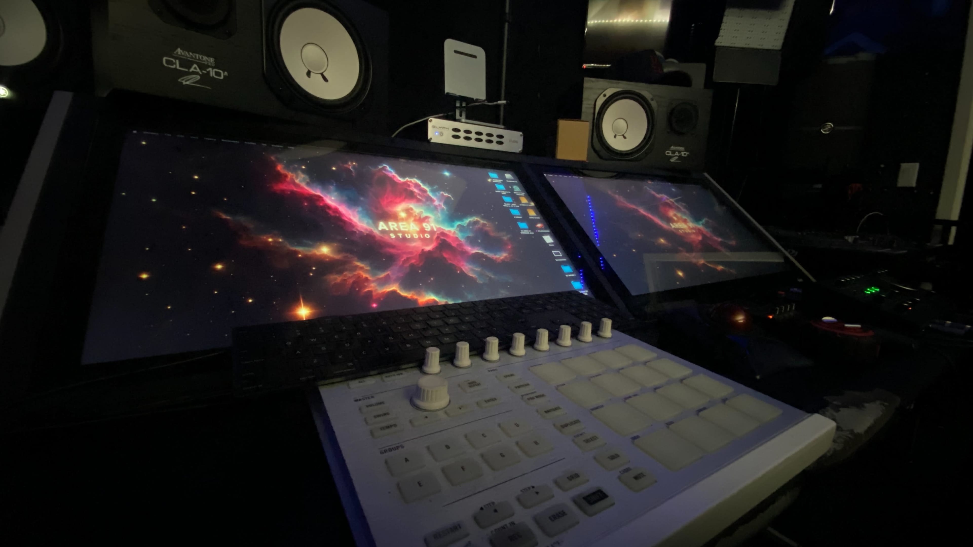 The image shows a music production setup featuring dual computer monitors displaying a cosmic background, a MIDI controller, and studio monitors in a dimly lit environment.