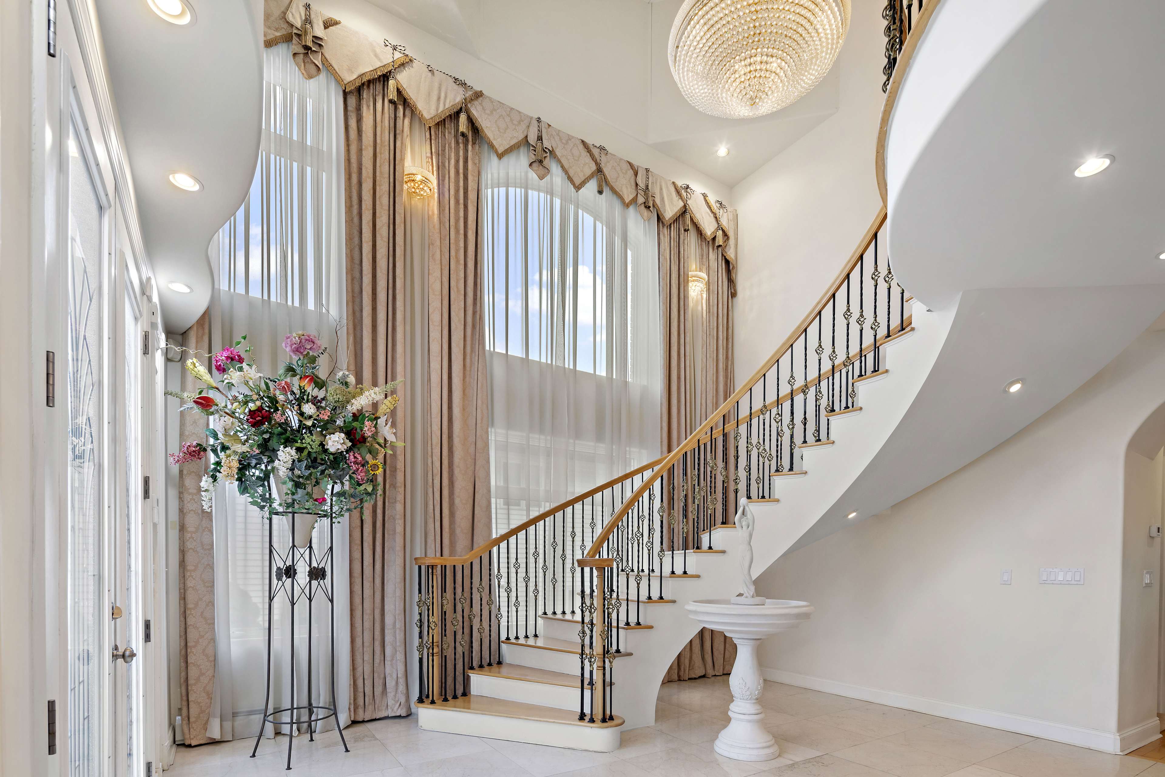 An elegant entryway with a sweeping staircase, large windows, draped curtains, and a floral arrangement on a stand.