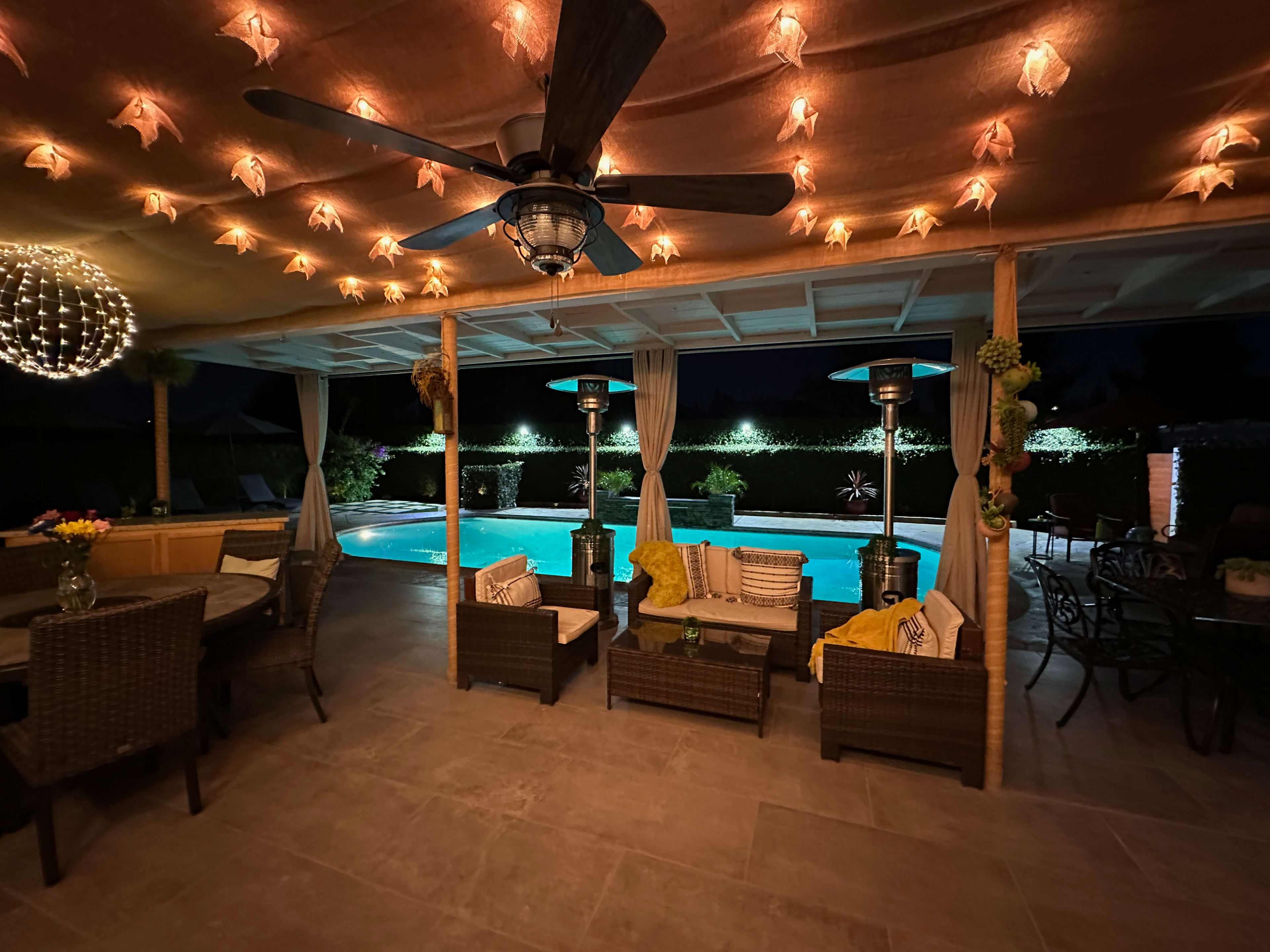 The image shows a well-lit outdoor patio area with wicker furniture, a ceiling fan, and a swimming pool illuminated by pool lights.