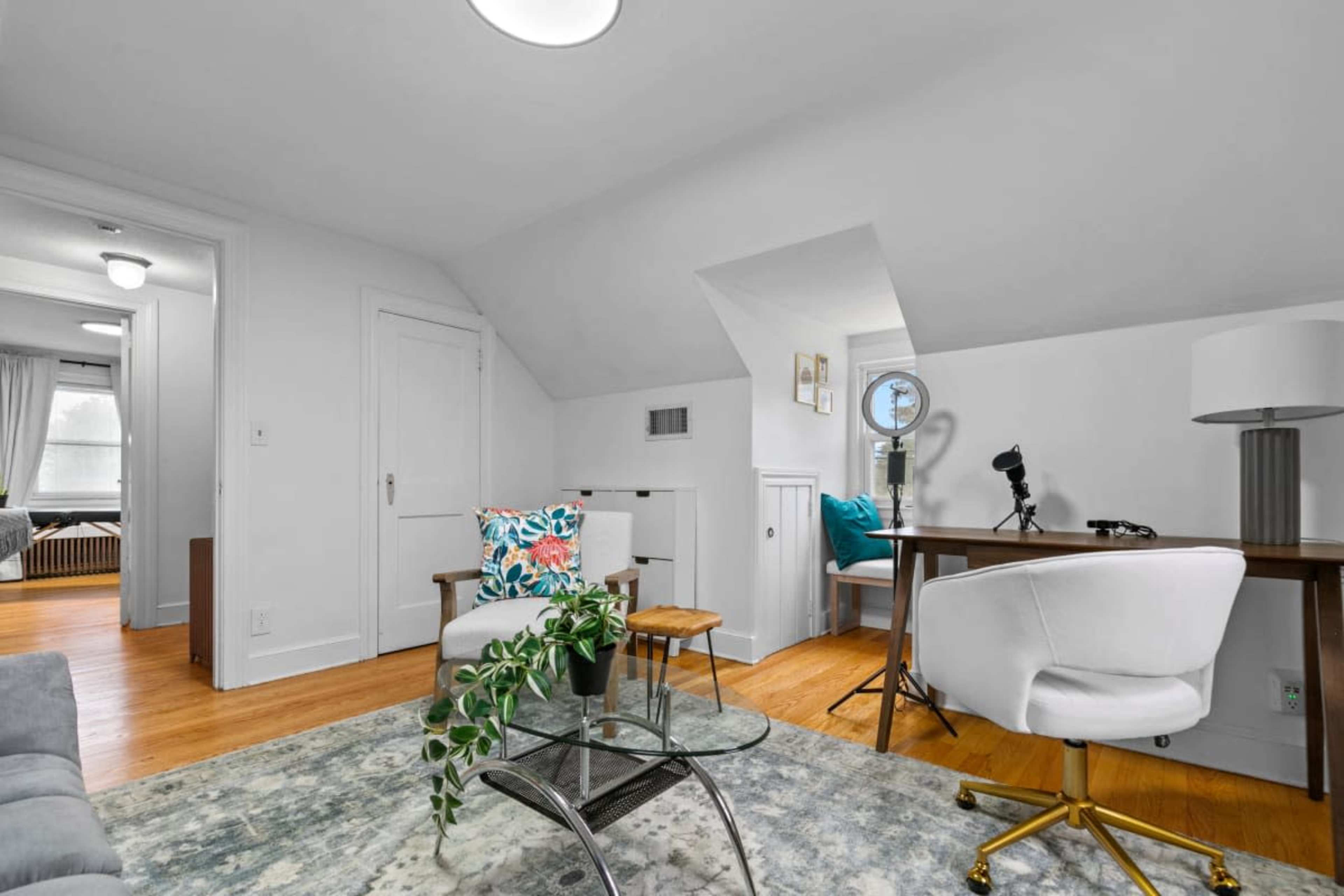 A light-filled attic room features a small seating area, a desk with a microphone, and a round glass coffee table on a patterned rug.