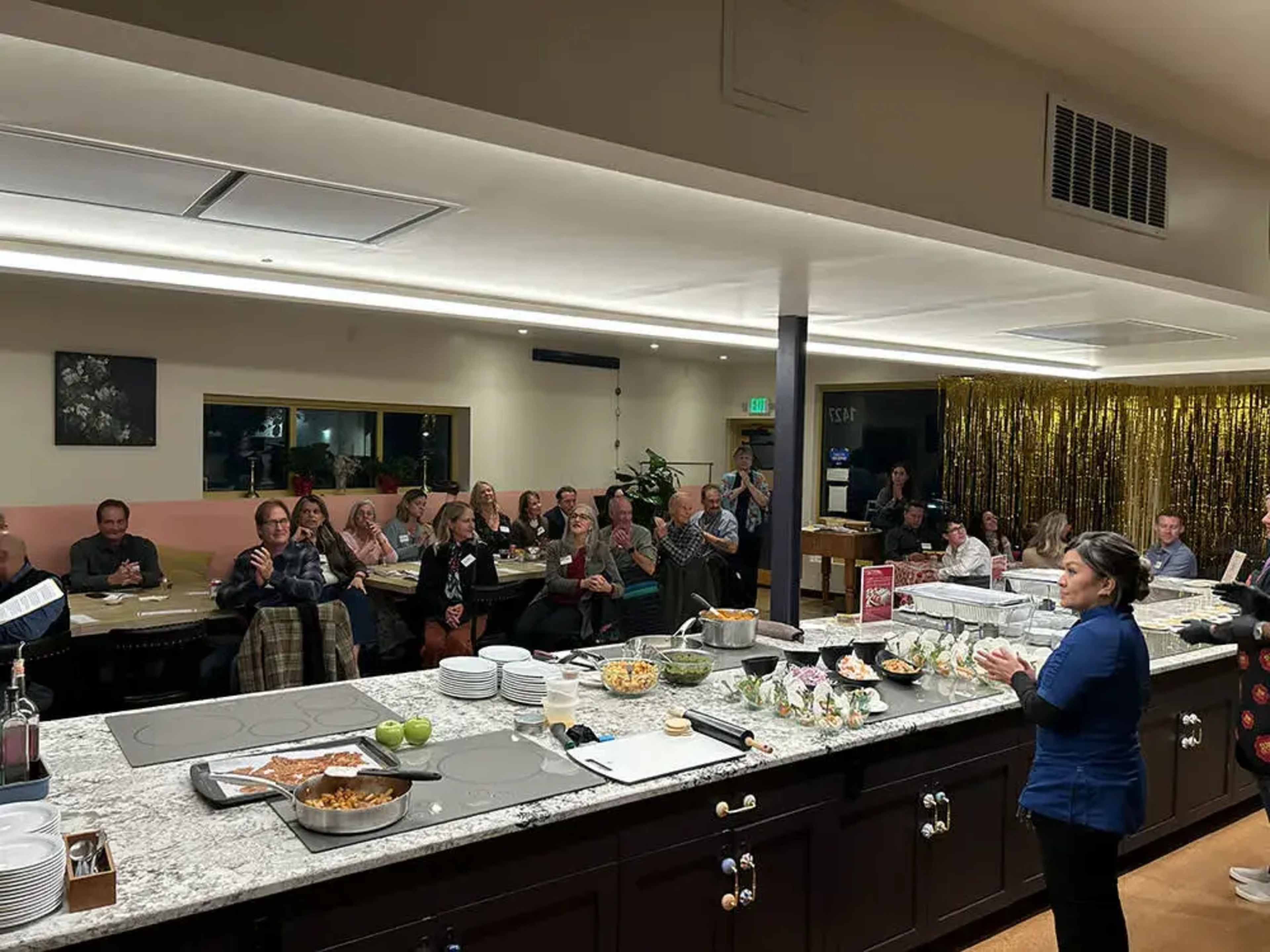 A woman stands at the front of a dining area, addressing an audience seated at tables filled with various dishes.