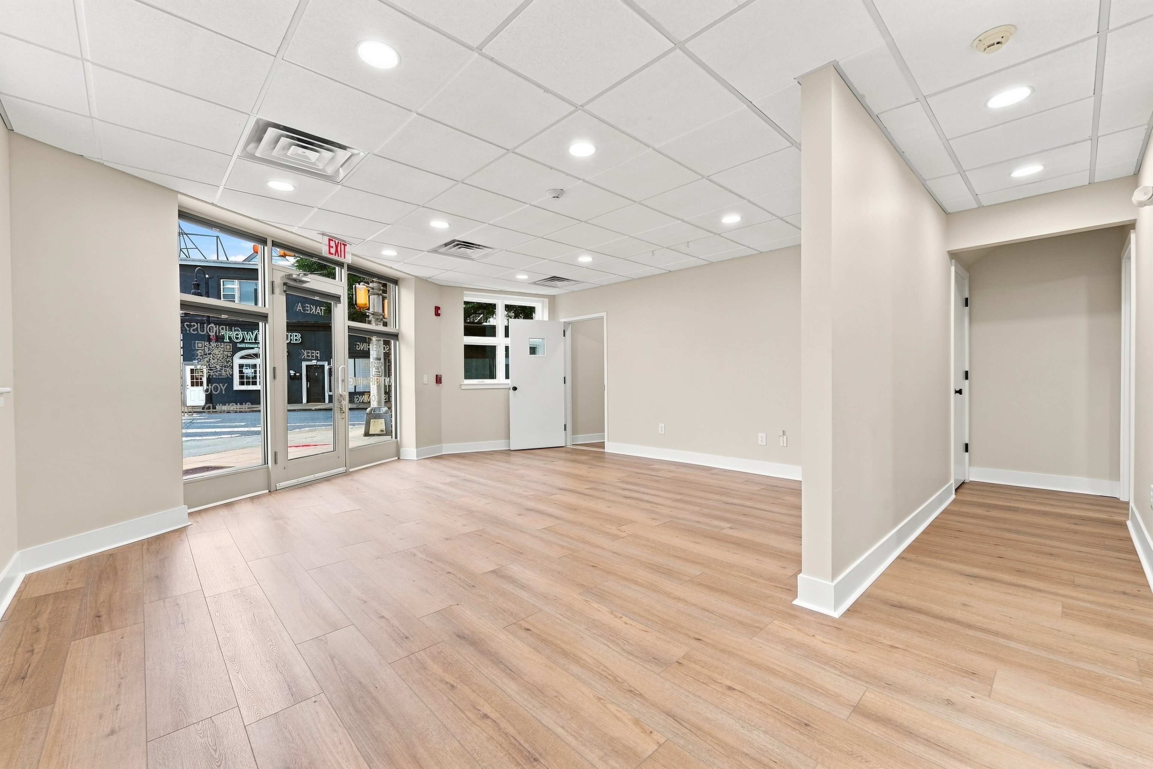 The image shows a vacant, well-lit interior space with light wood flooring and a large entrance area featuring glass doors.