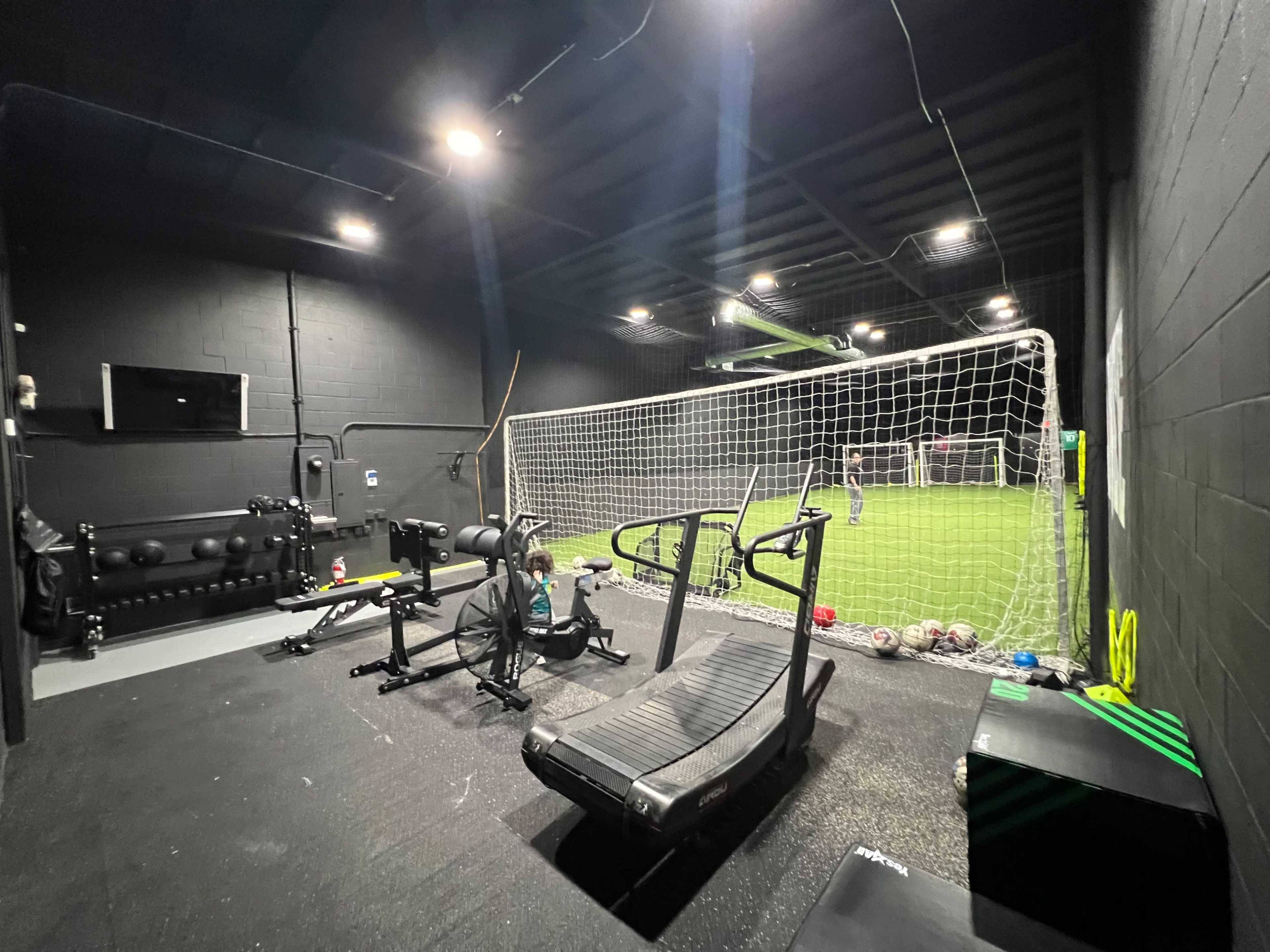 A dark training room with fitness equipment and a soccer goal in the background.