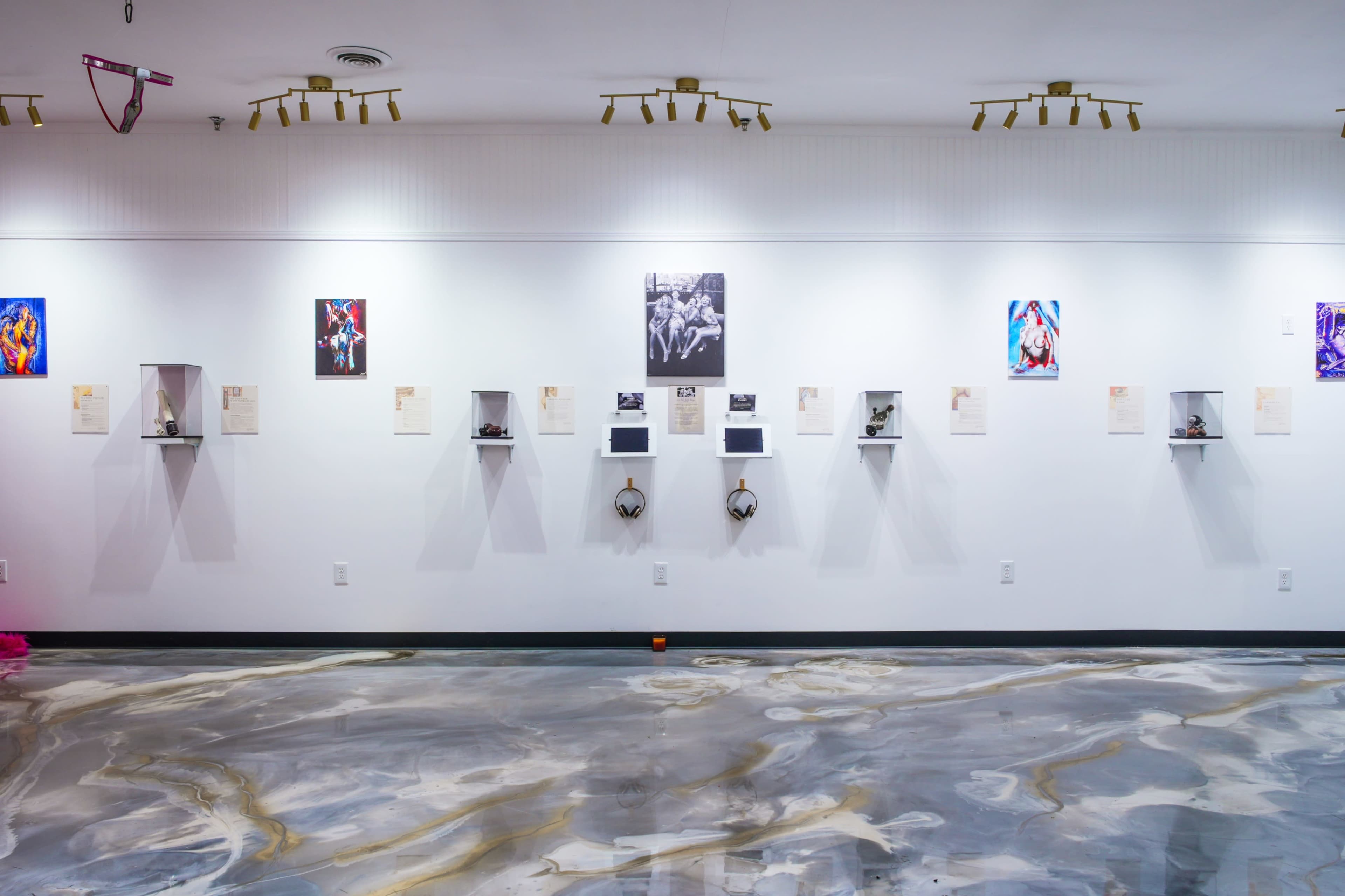 The image shows a gallery wall with various framed artworks displayed on shelves against a smooth, marbled floor.