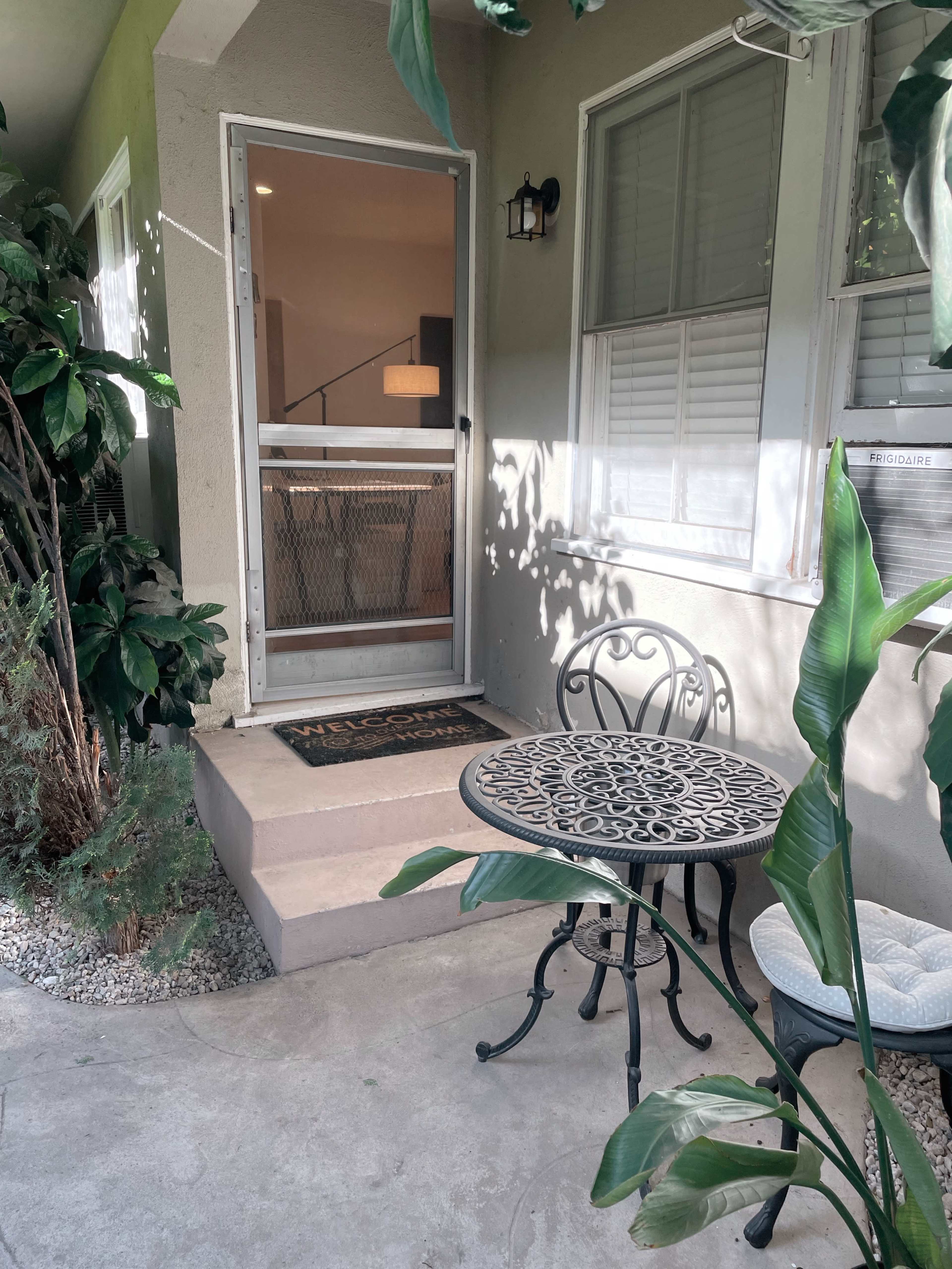 A small outdoor space features a black metal table and chair set, surrounded by plants, with a door leading into a house.