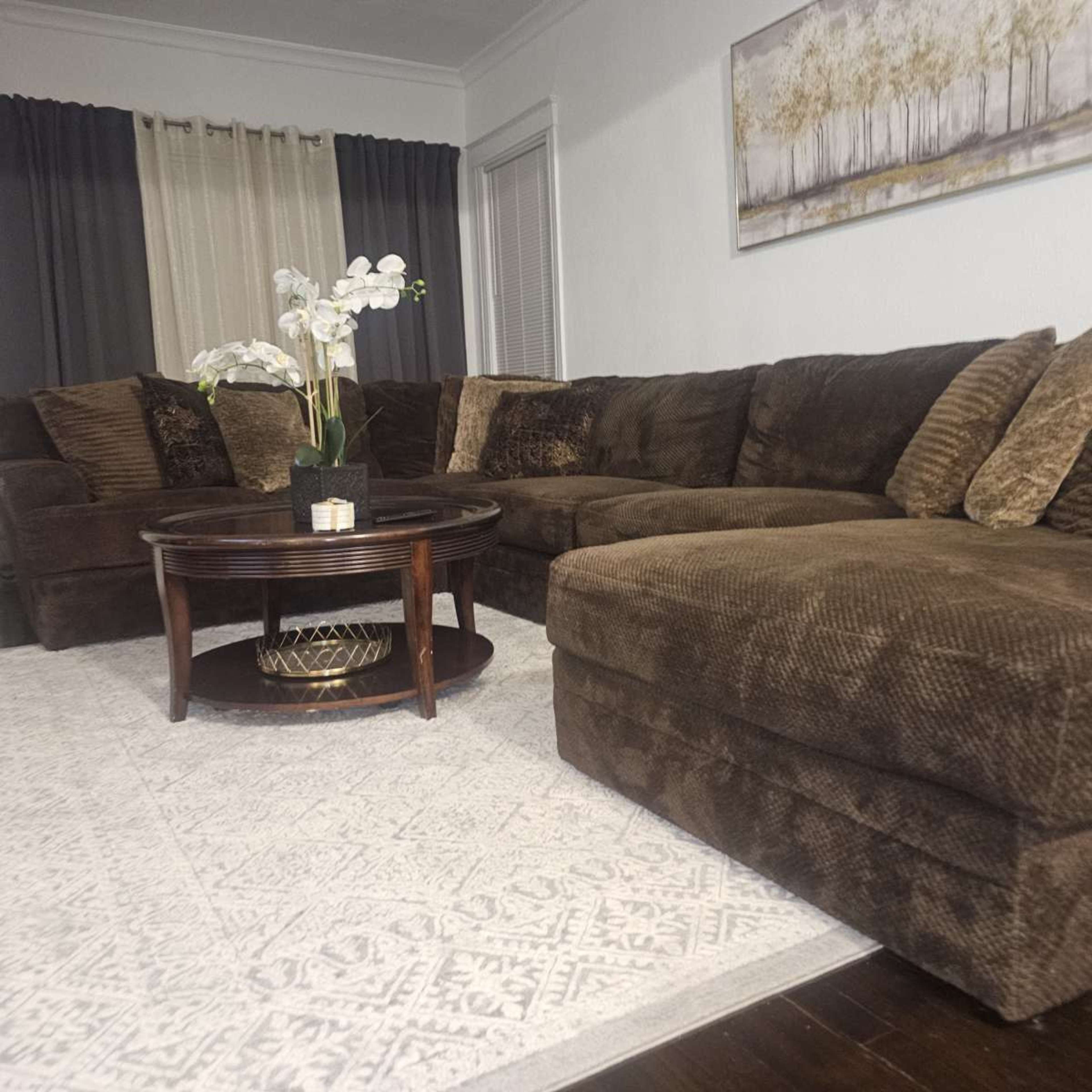 A sectional sofa with various cushions is positioned in a cozy living room, accompanied by a round coffee table and decorative plants.