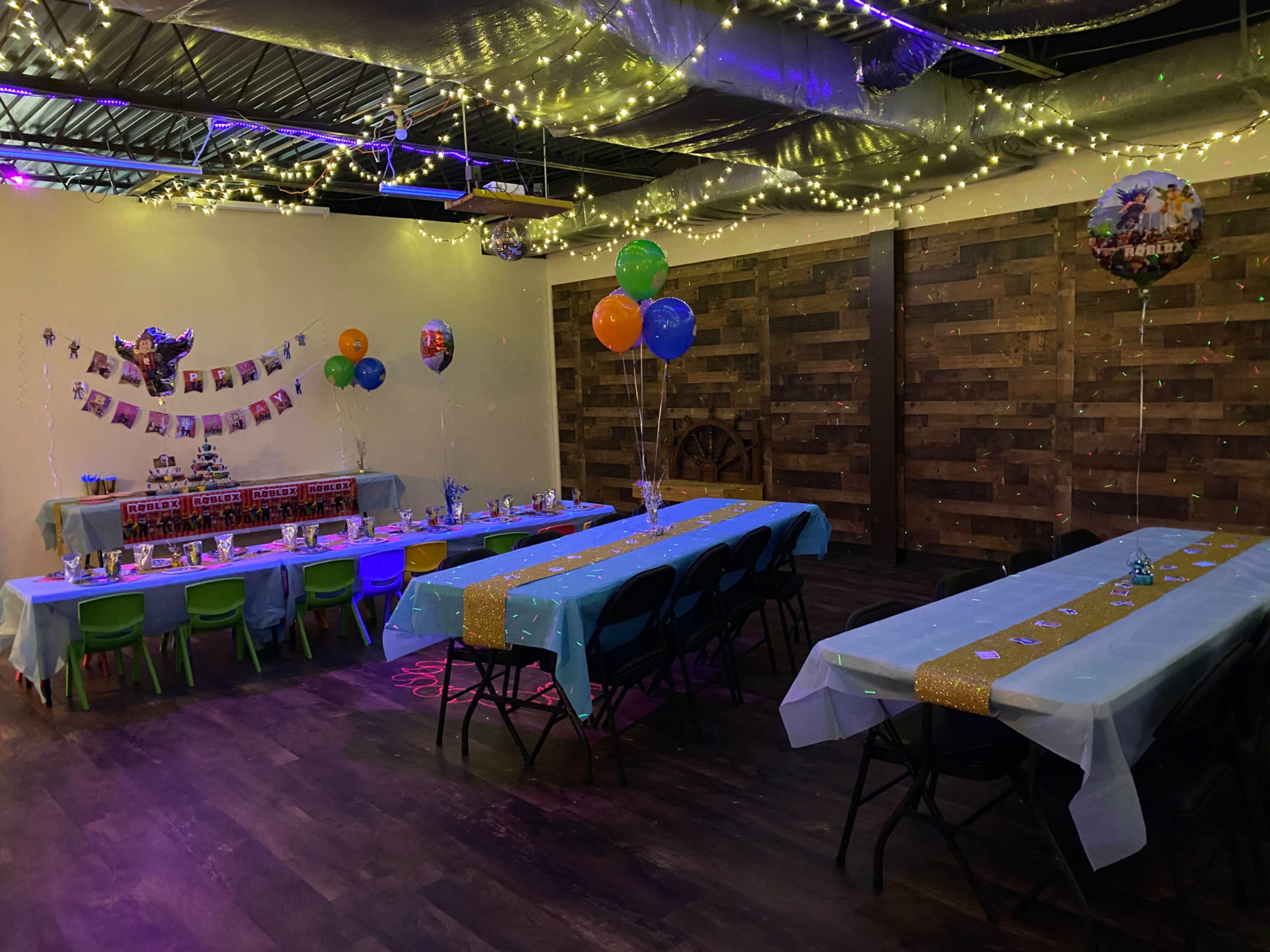 A decorated party venue features multiple tables with blue tablecloths, colorful balloons, and string lights overhead.