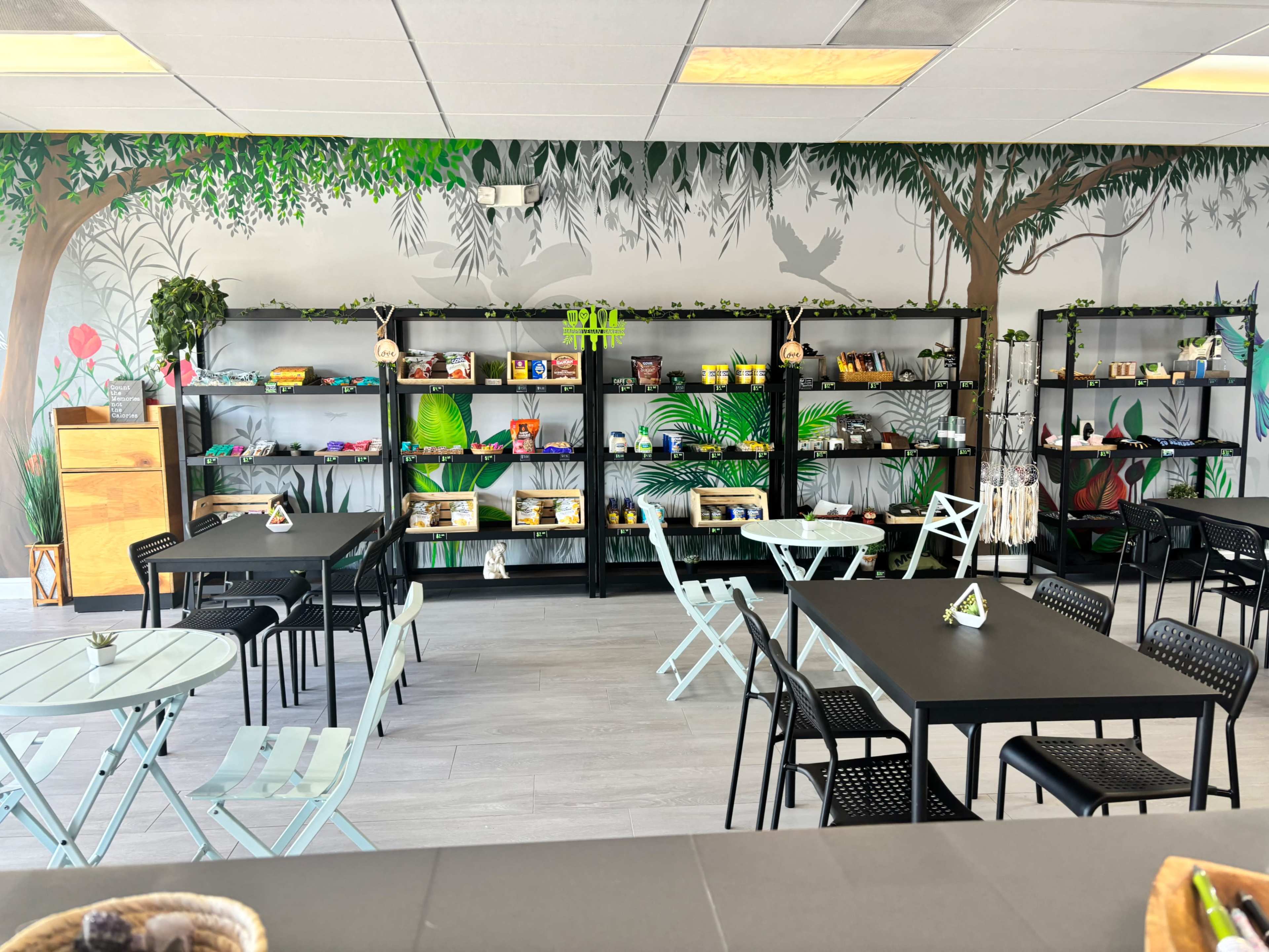 A brightly lit café features black tables and chairs arranged around green-painted walls adorned with tropical murals and shelves filled with various items.