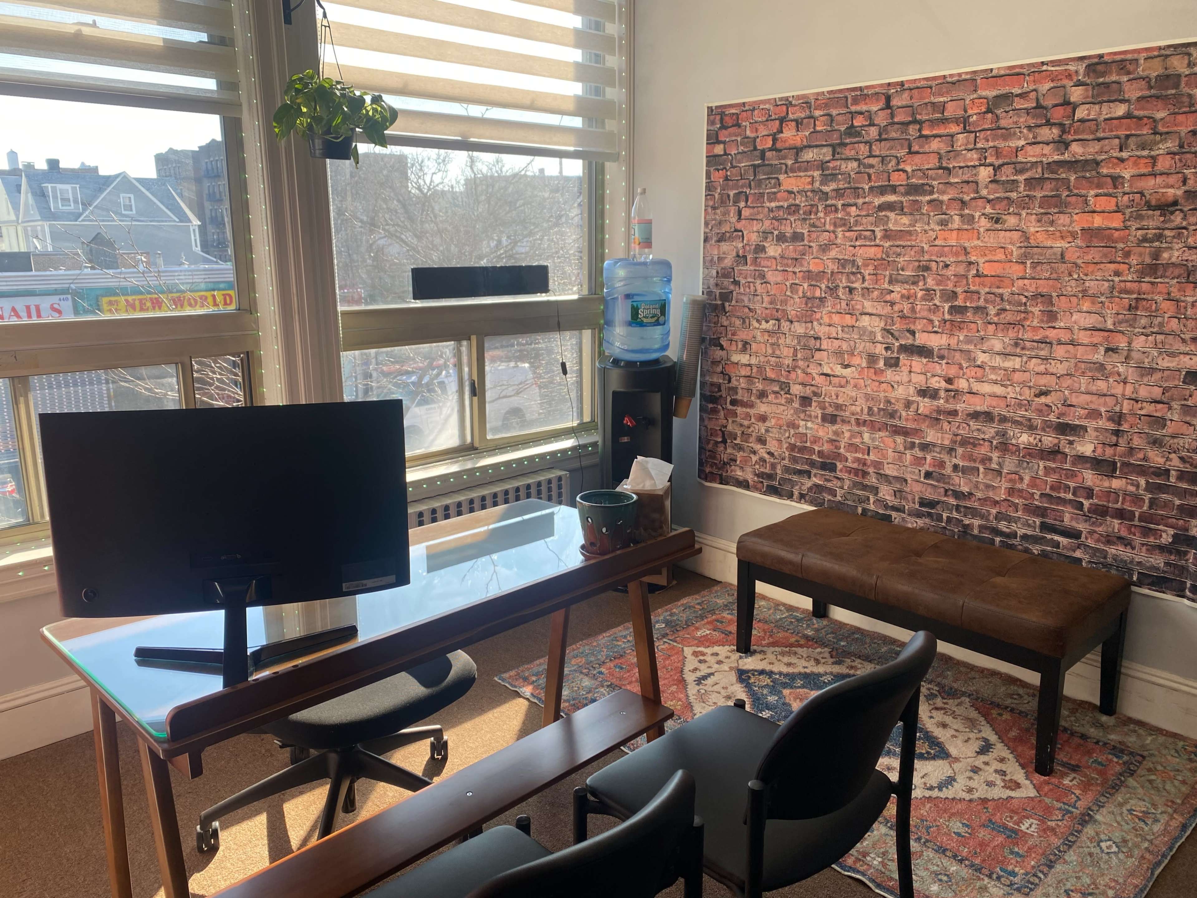 An office space with a glass desk, a monitor, a water cooler, and a bench along a wall featuring a brick pattern.