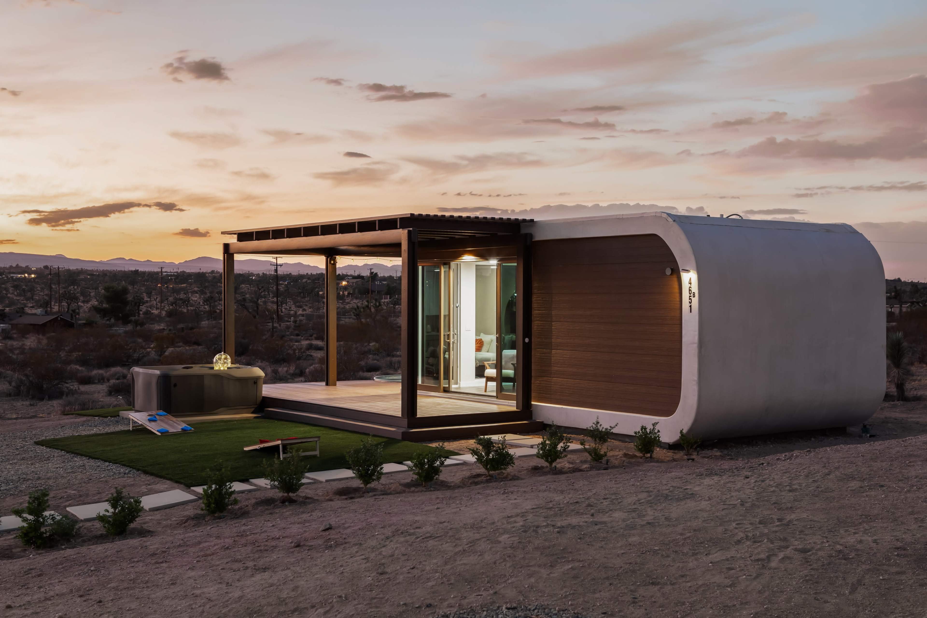 A modern, curved tiny home features large glass sliding doors, wooden accents, and is set against a desert landscape at sunset.