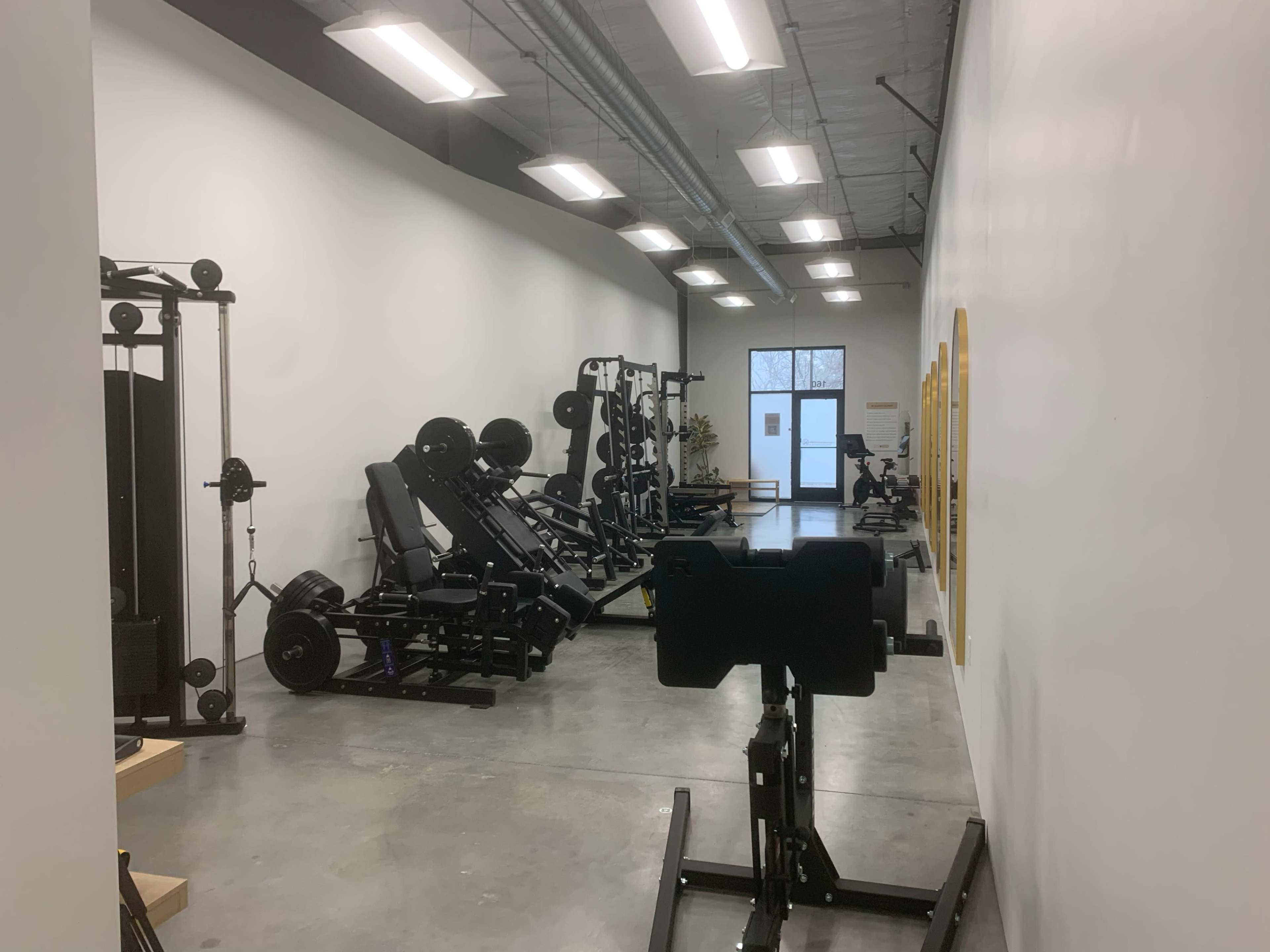 The image shows a fitness facility with various gym equipment arranged along the walls and a large window at the far end.