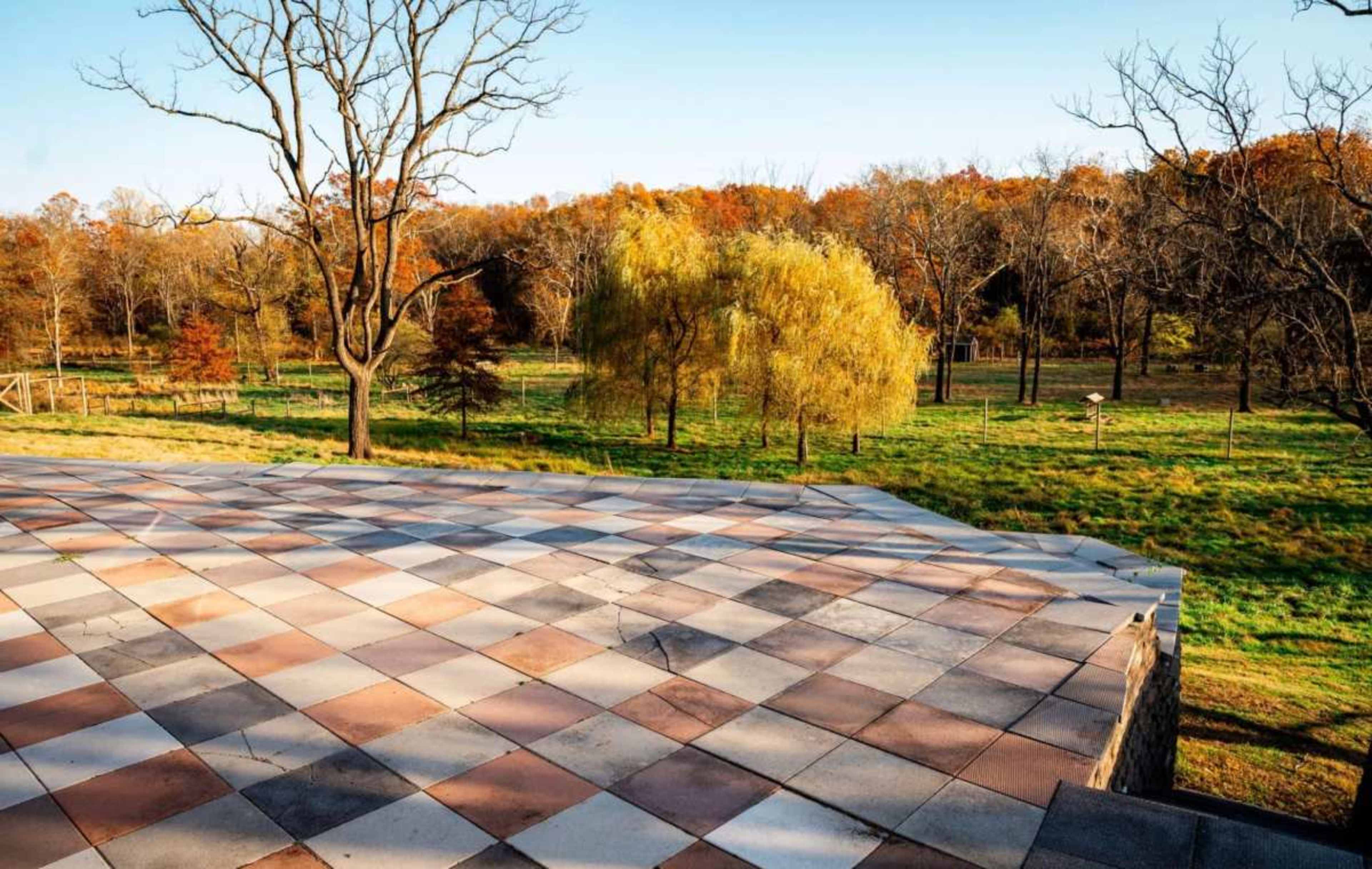 A patterned stone terrace overlooks a field with trees and autumn foliage in the background.