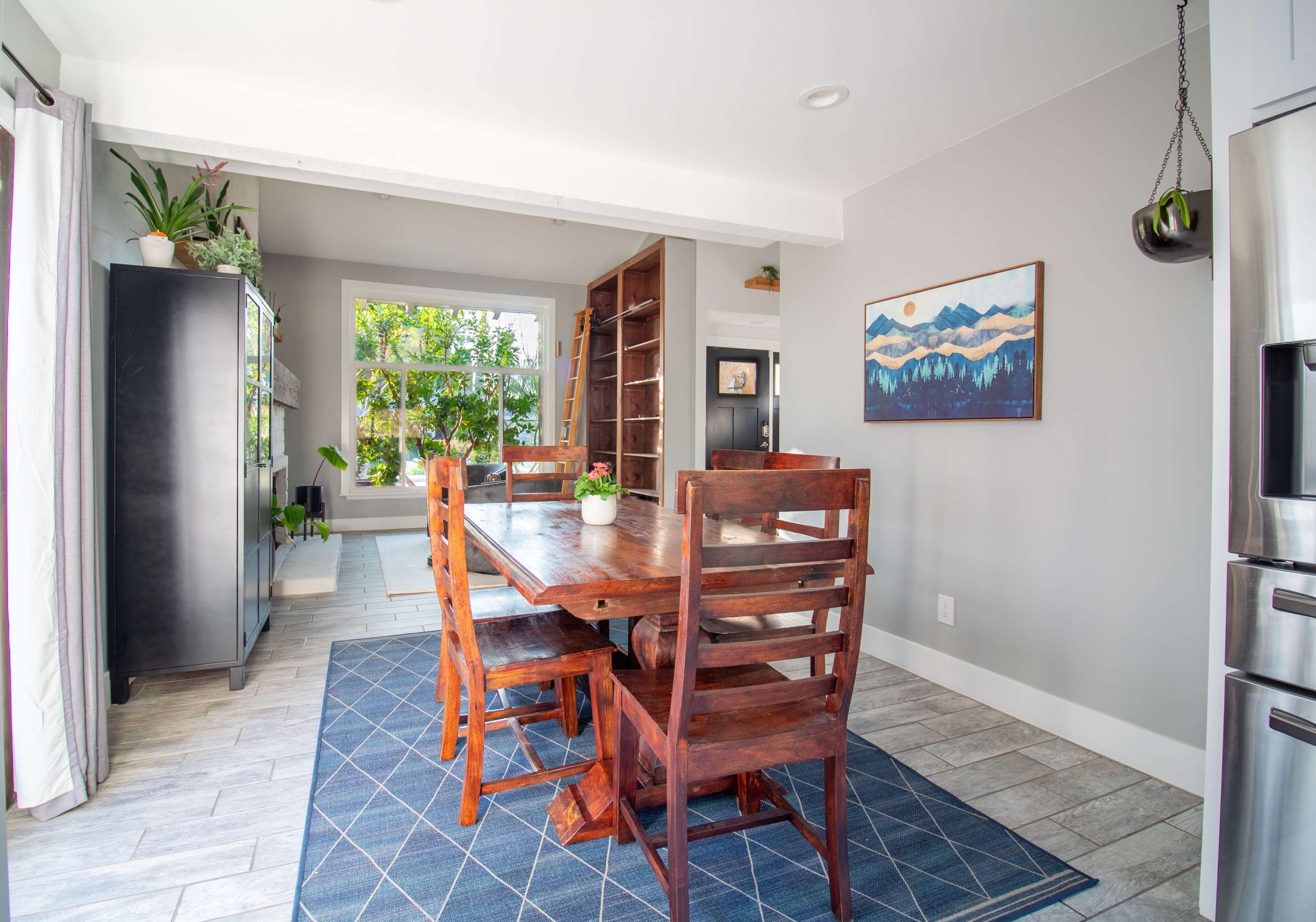 A wooden dining table with four chairs is positioned in a well-lit room that features large windows and a bookshelf against the wall.