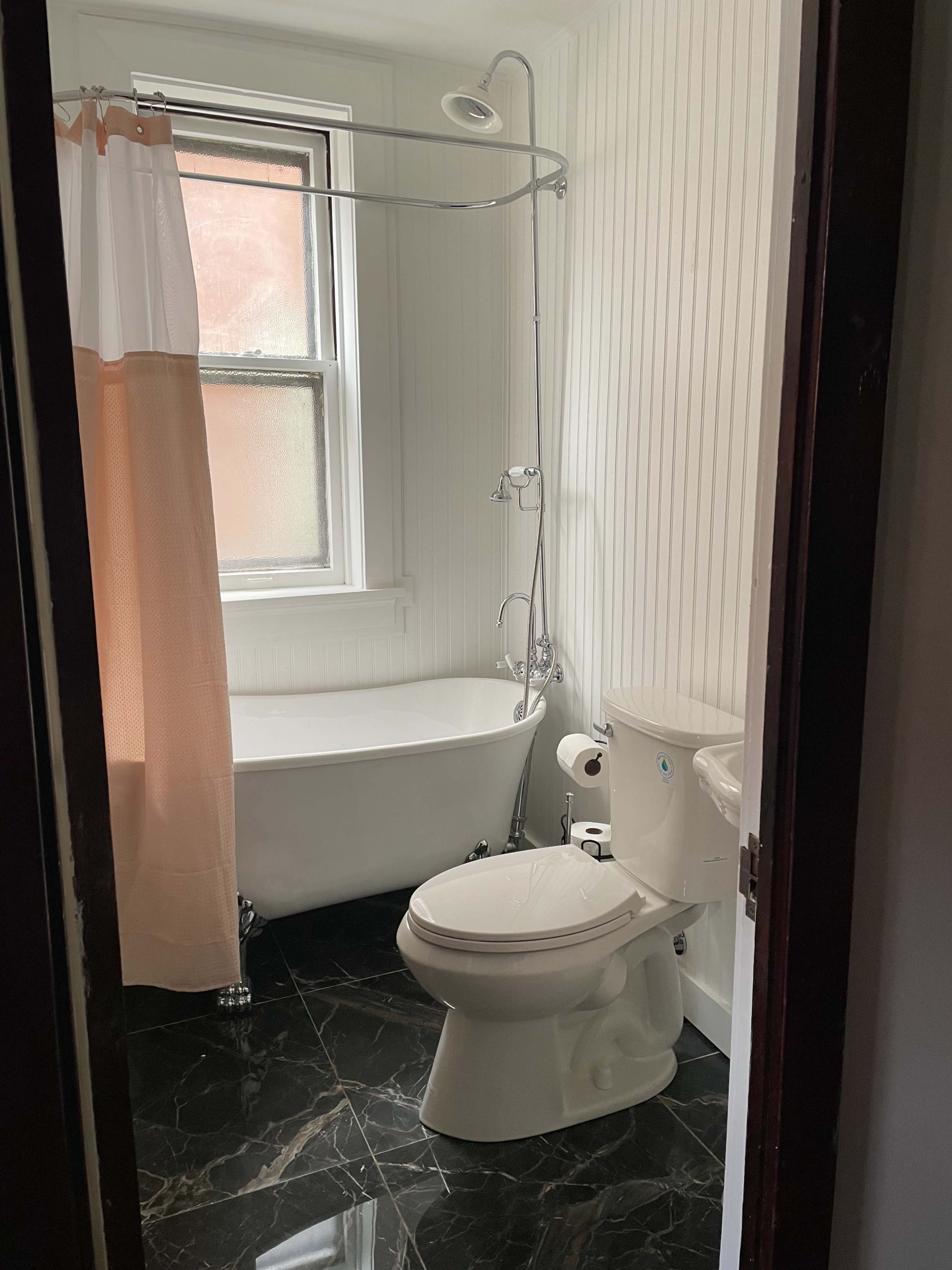 A small bathroom featuring a clawfoot bathtub, a shower attachment, a toilet, and light-colored walls with a contrasting dark floor.
