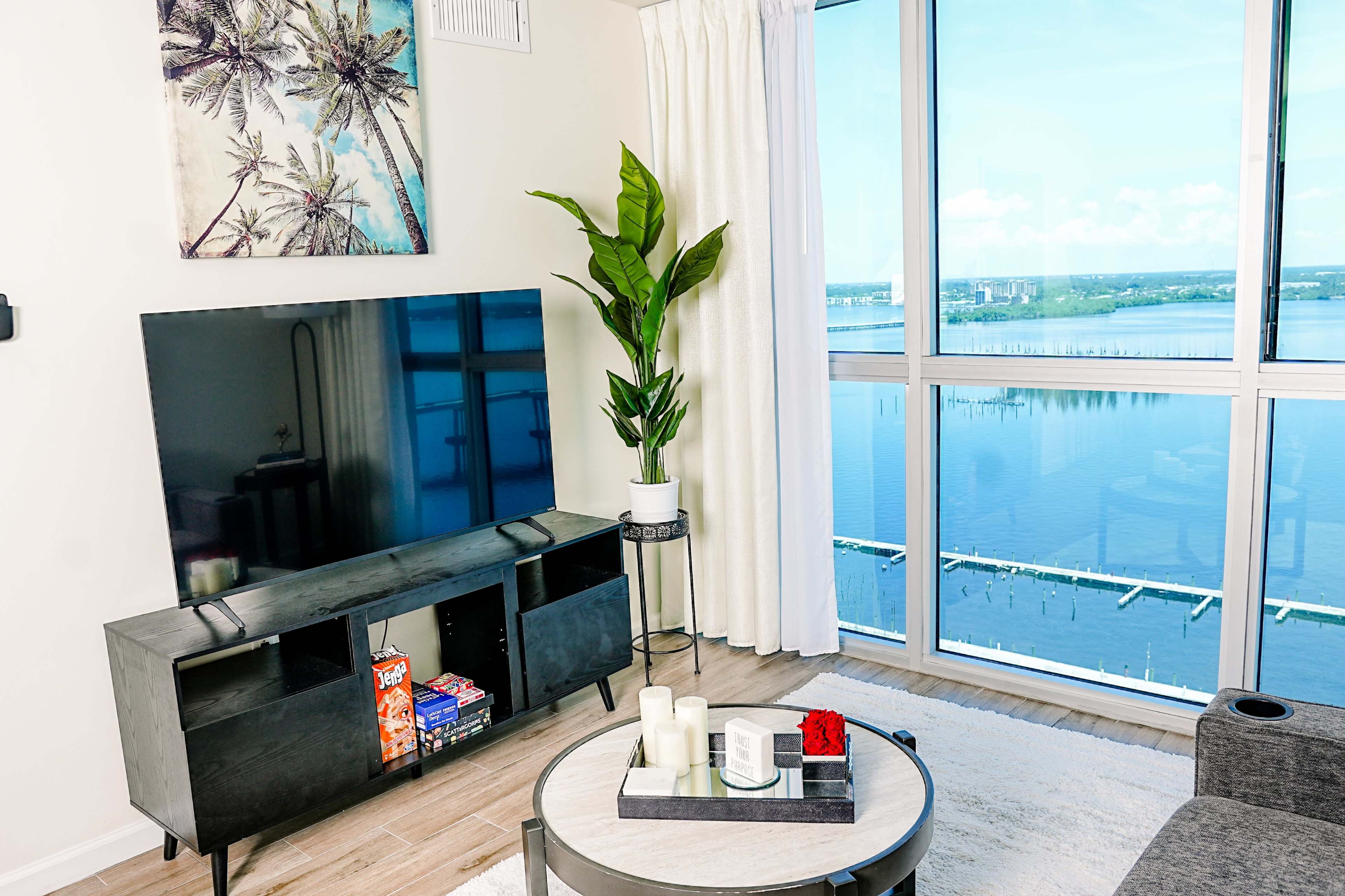 A contemporary living room features a large television on a sleek black cabinet, a potted plant, and a view of water through floor-to-ceiling windows.