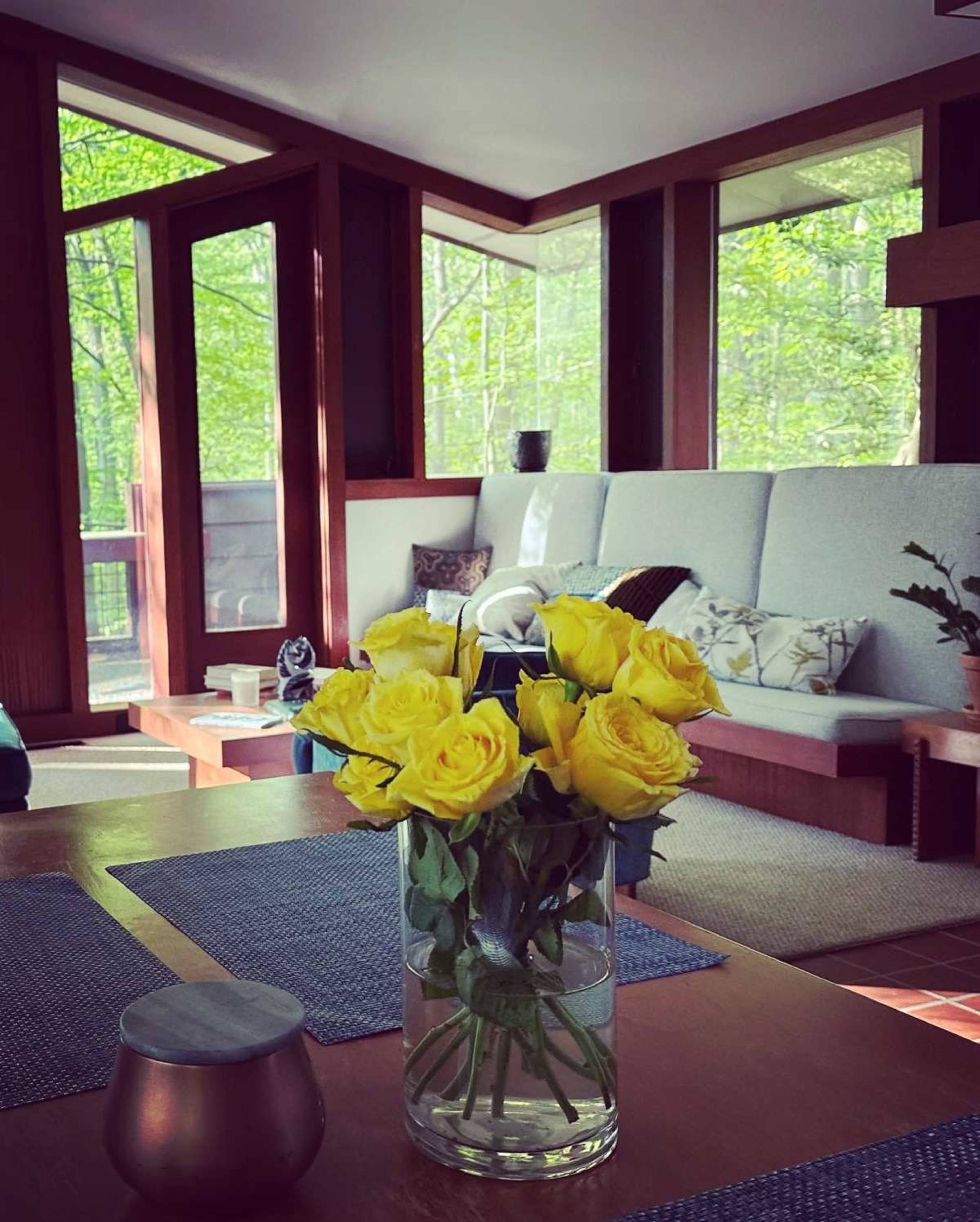 A vase of yellow roses sits on a wooden table in a living room featuring a light-colored sofa and large windows overlooking a forested area.