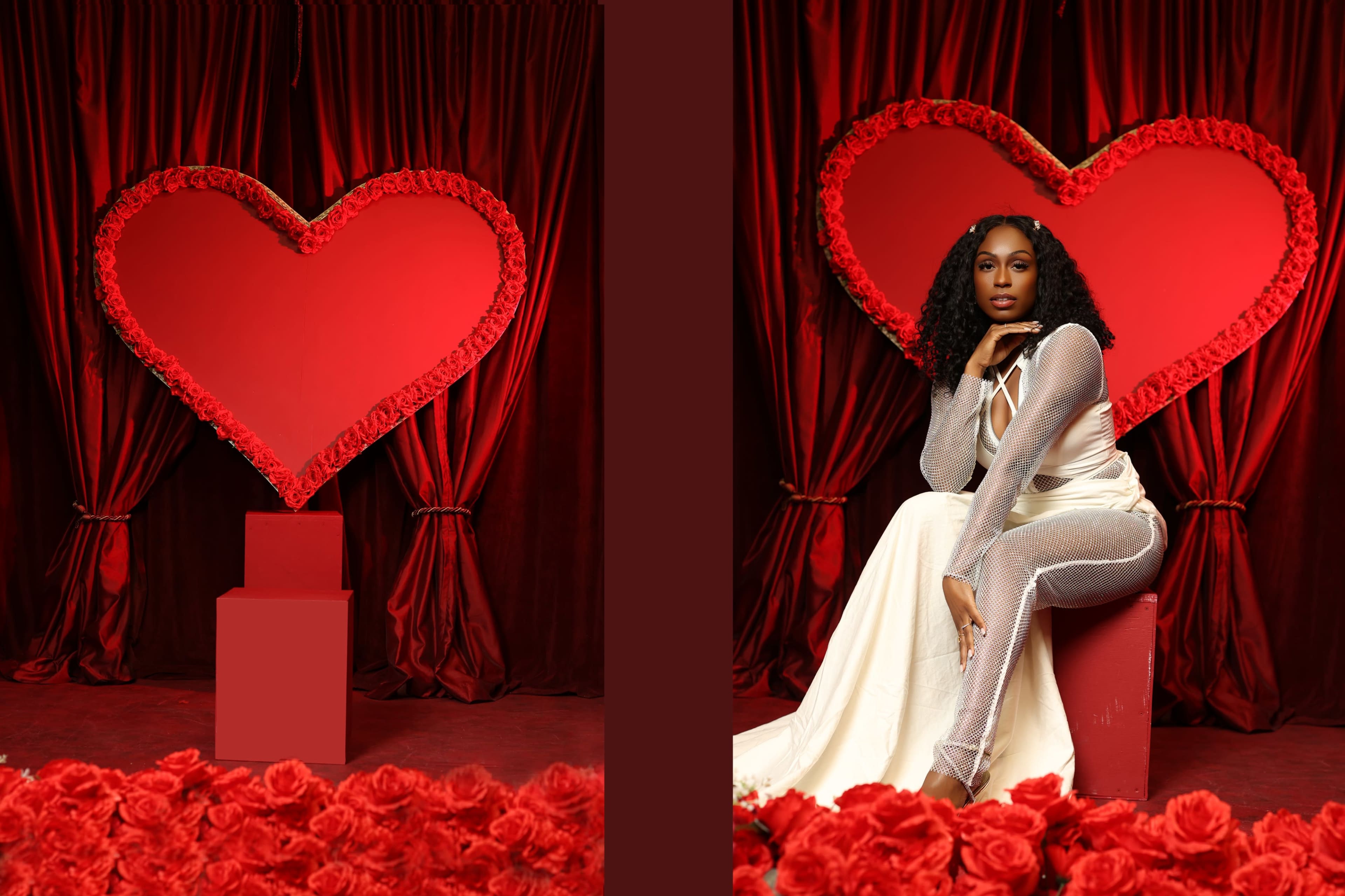 A woman dressed in a flowing white gown poses next to a large heart-shaped display made of roses against a red backdrop.