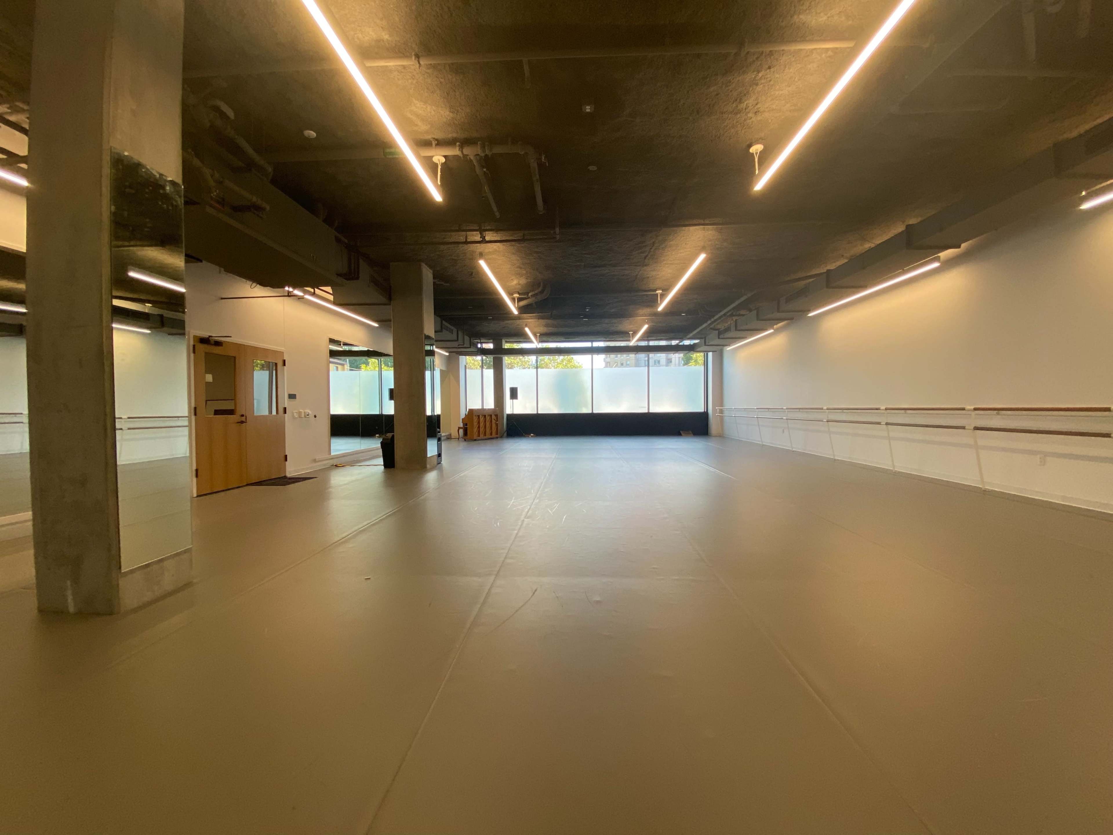 A spacious dance studio with a smooth floor, large windows, and wooden barre along the walls.