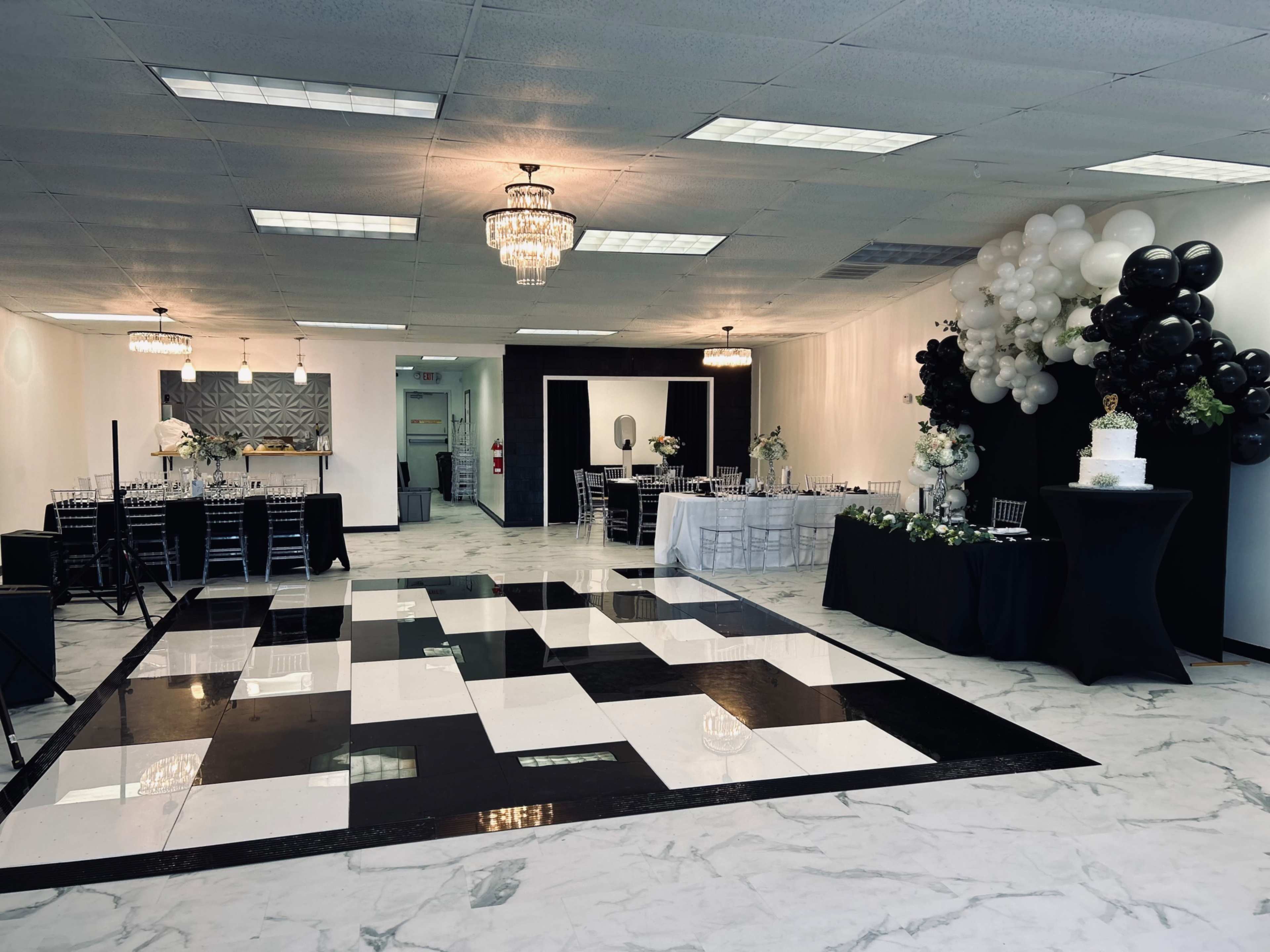 The image shows a decorated event space featuring a black and white checkered dance floor, tables set for guests, and elegant balloon arrangements.