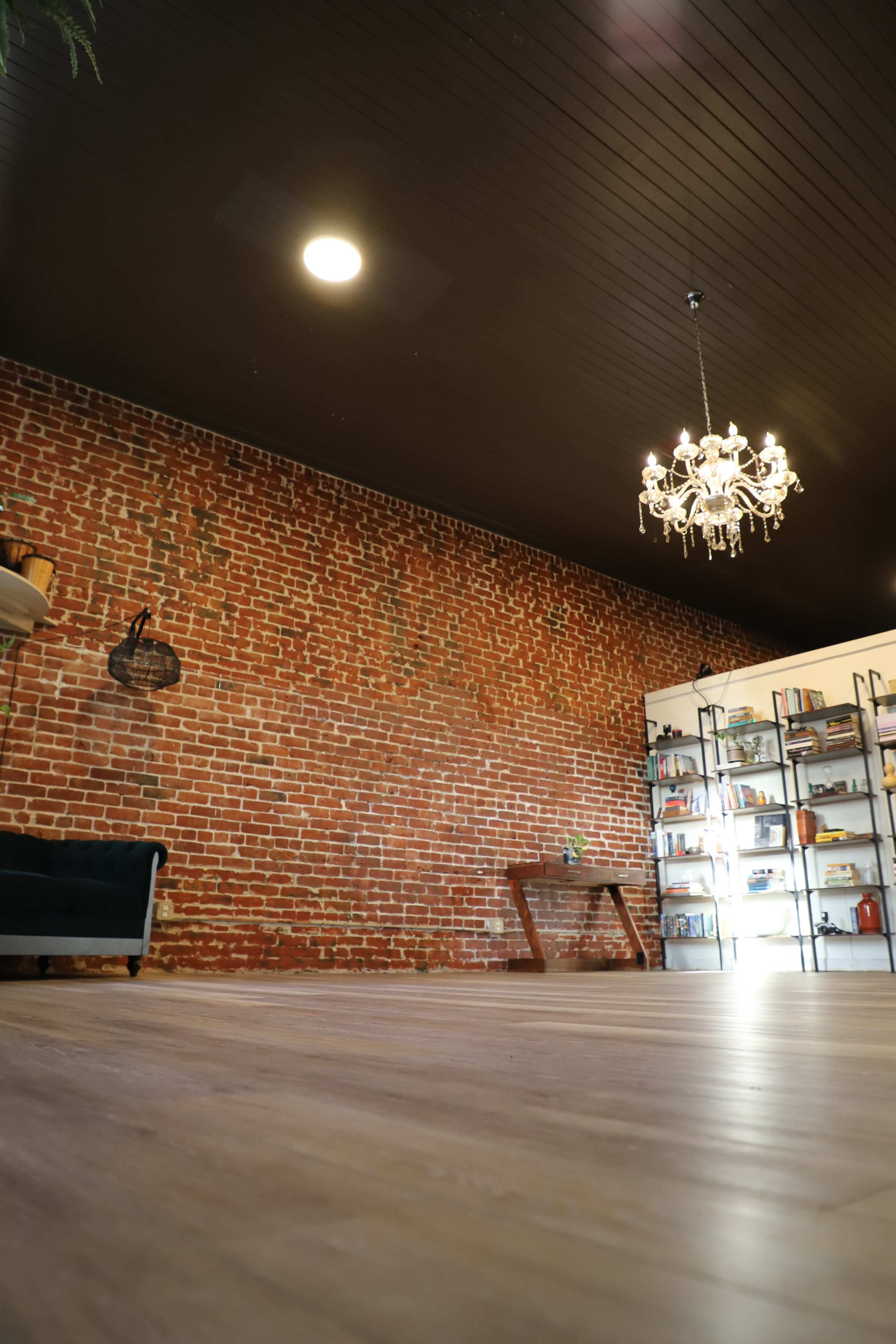 The image shows a spacious room with exposed brick walls, a wooden floor, and a chandelier hanging from the ceiling, alongside a bookshelf filled with various items.