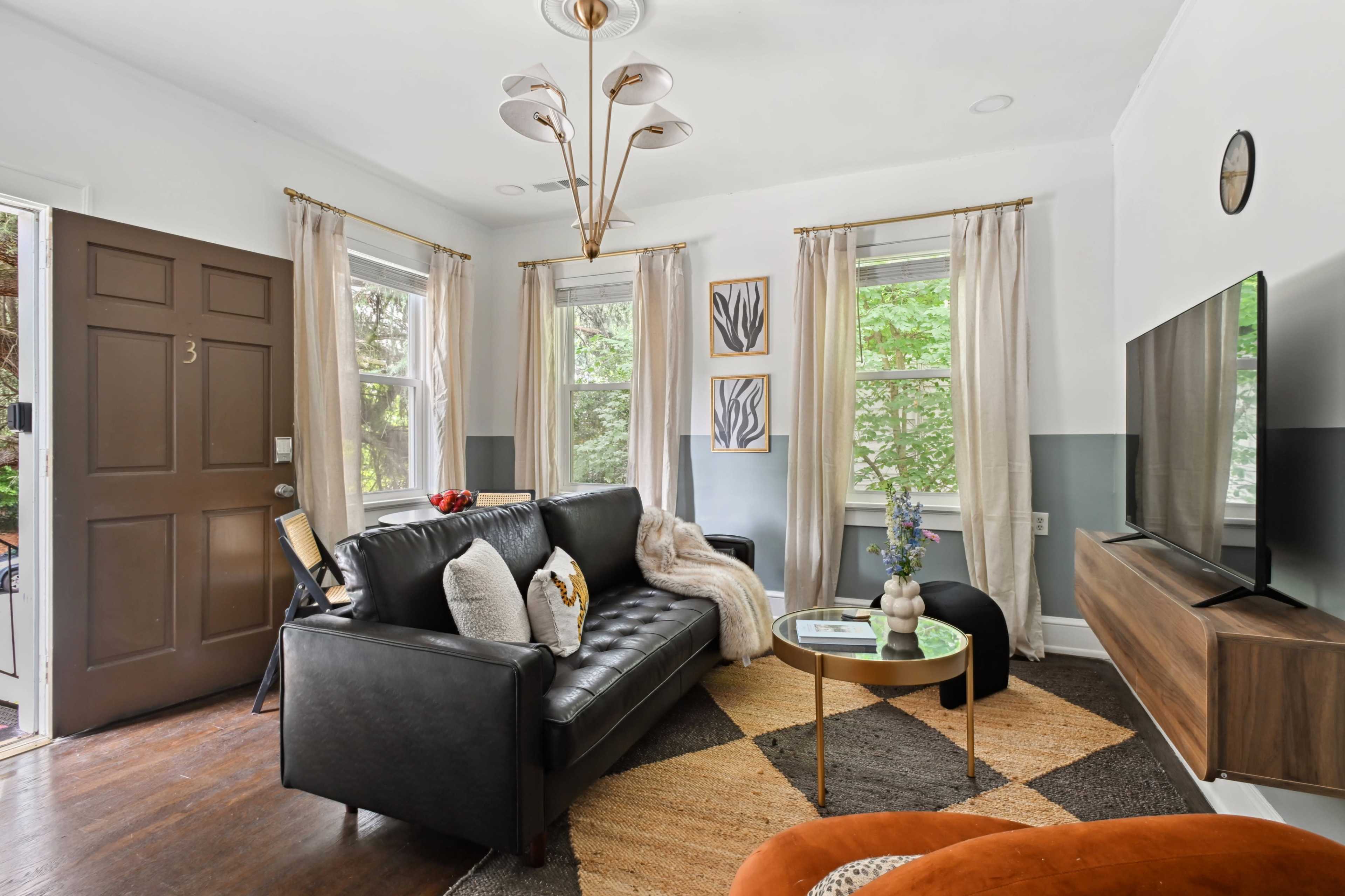 The living room features a black leather sofa, a round coffee table, and large windows with sheer curtains, creating a cozy atmosphere.