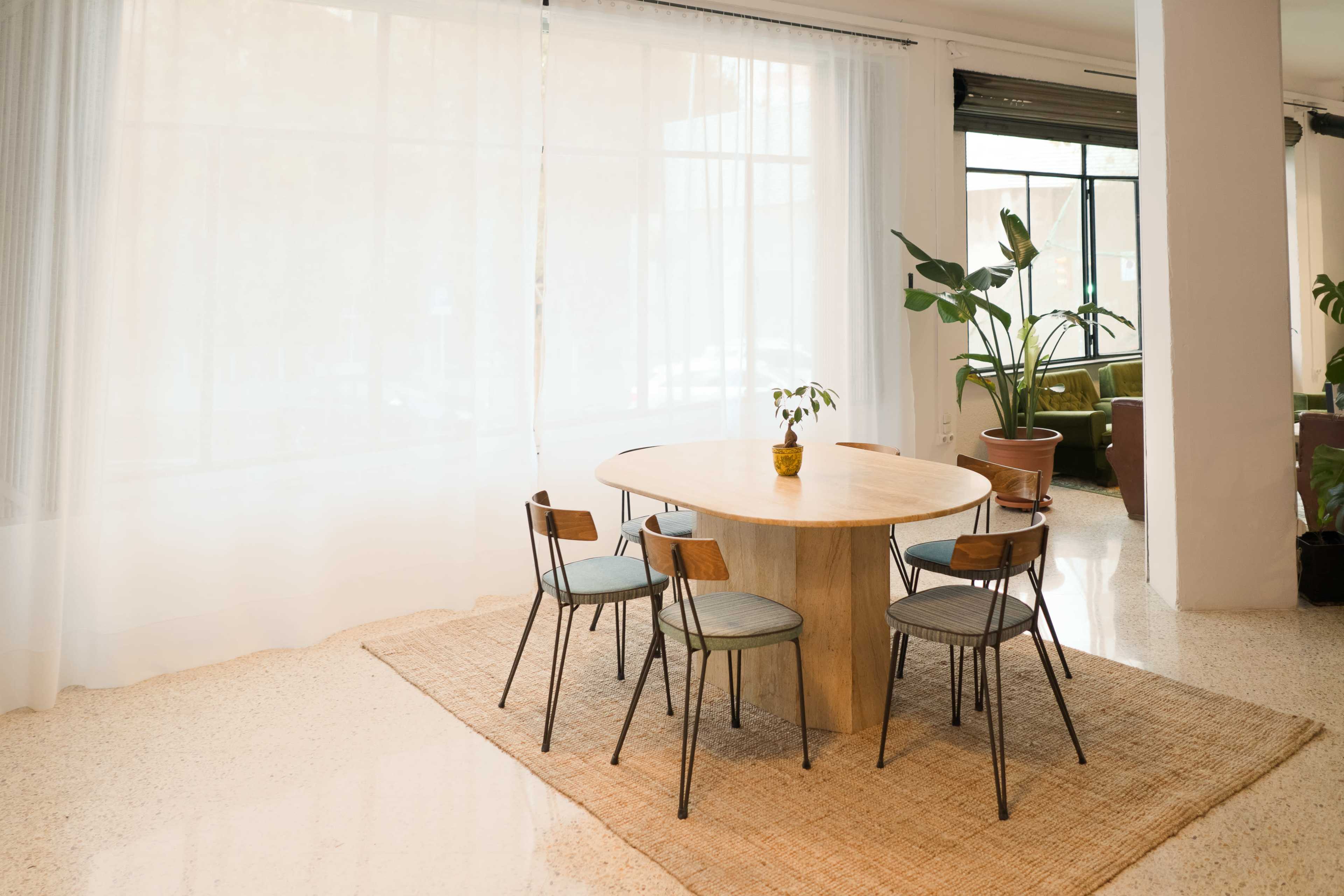 A round wooden table surrounded by four chairs sits on a woven rug near large windows with sheer curtains, accompanied by potted plants in the background.