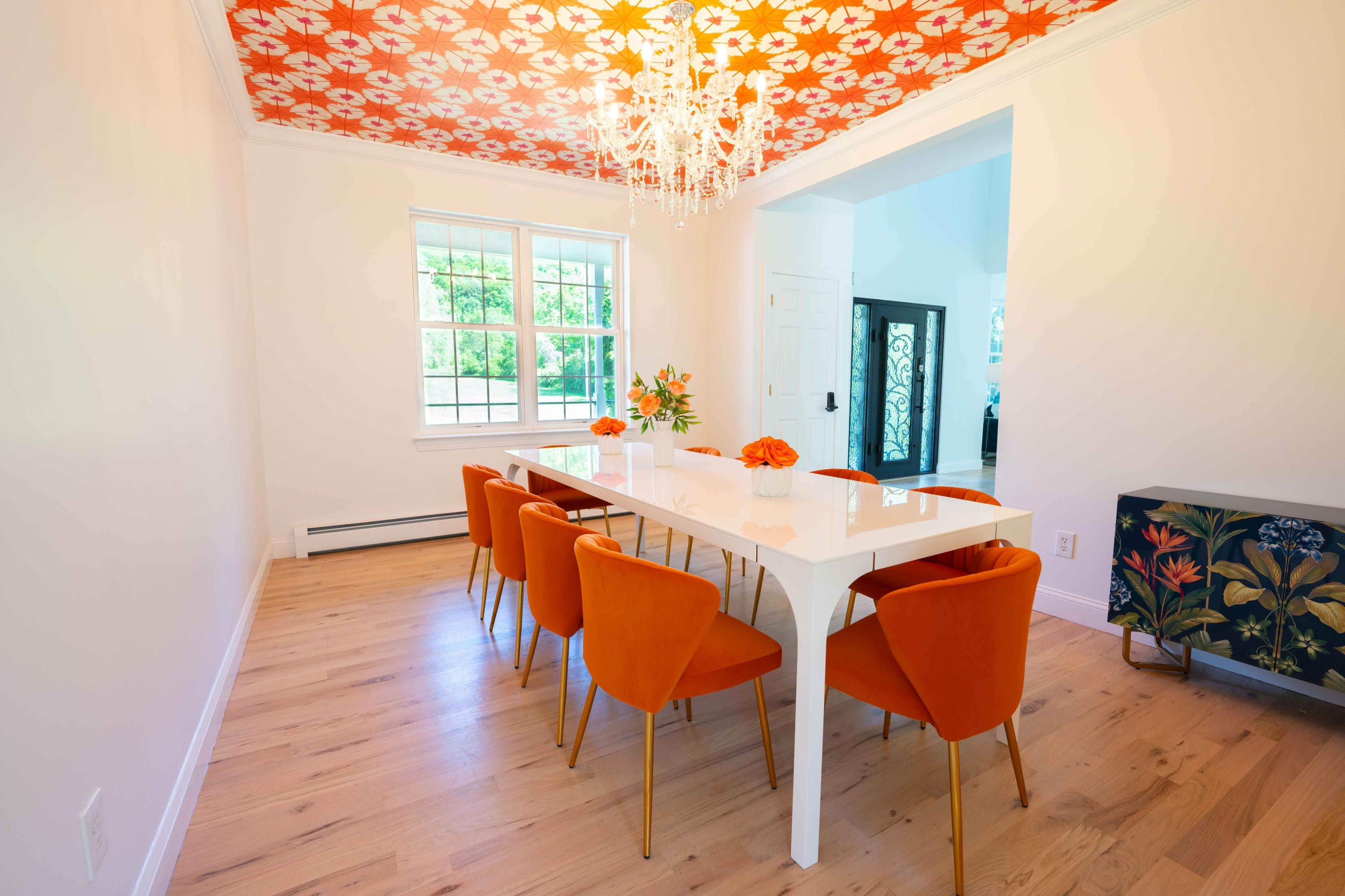 A modern dining room features a long white table surrounded by orange chairs, with a floral-patterned ceiling and a chandelier above.