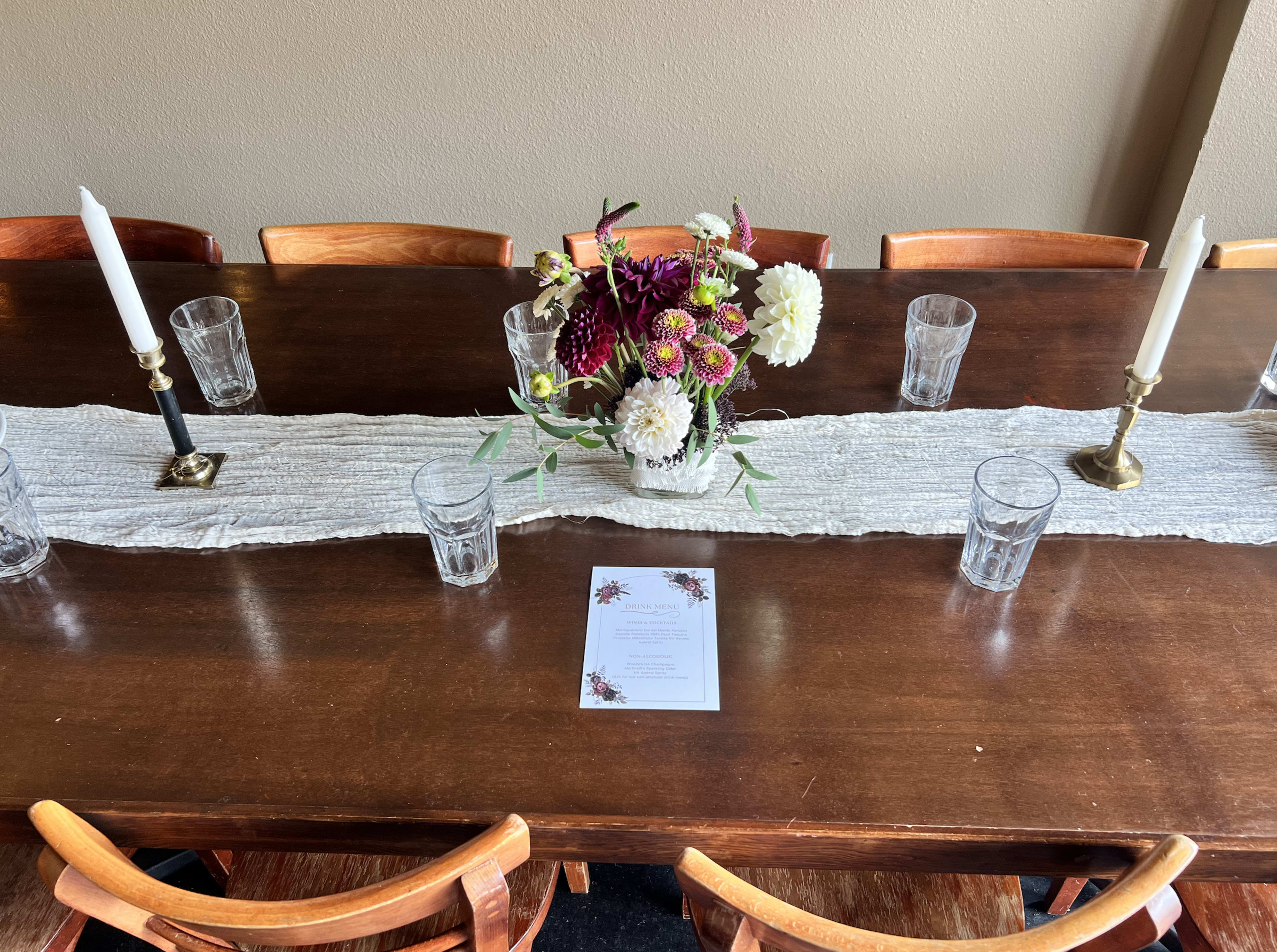 A long wooden dining table is decorated with a floral centerpiece, lit candles, and glasses, accompanied by a menu placed on a linen runner.
