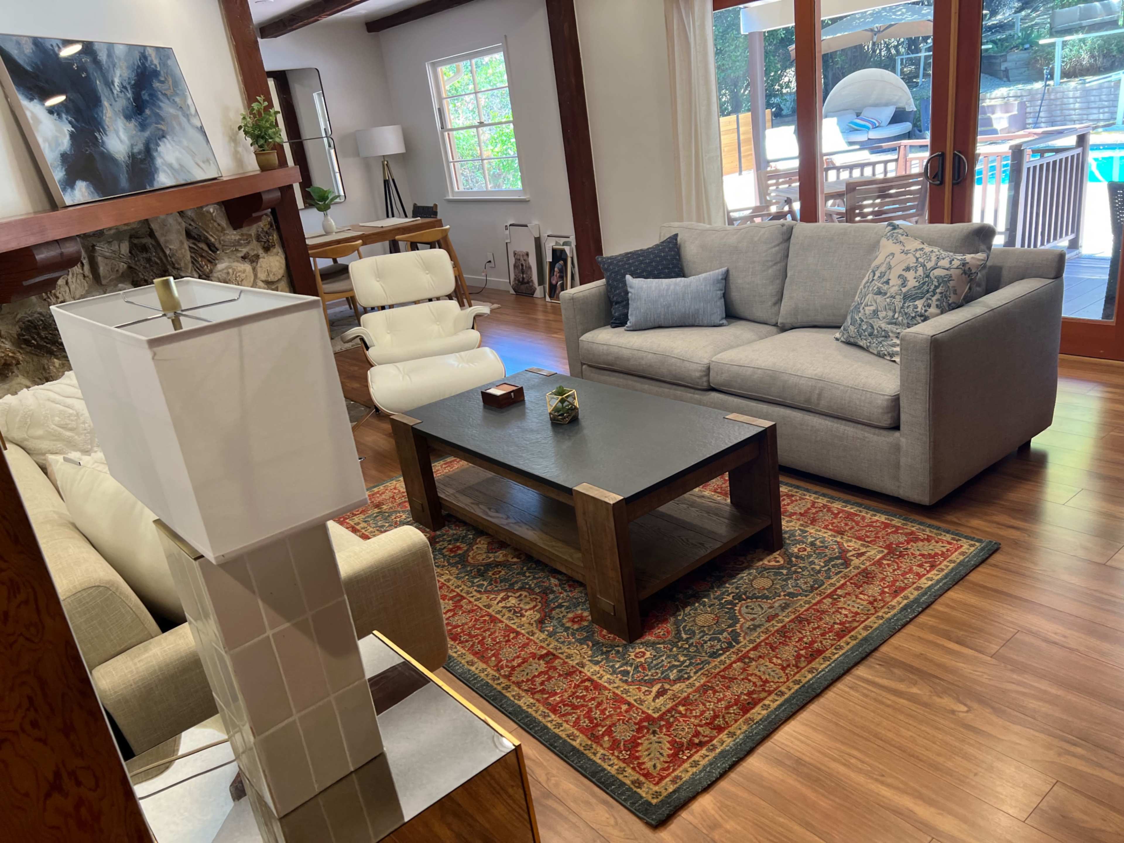 The living room features a gray sofa and an armchair surrounding a wooden coffee table, with a rug underneath and large windows overlooking a patio and pool area.