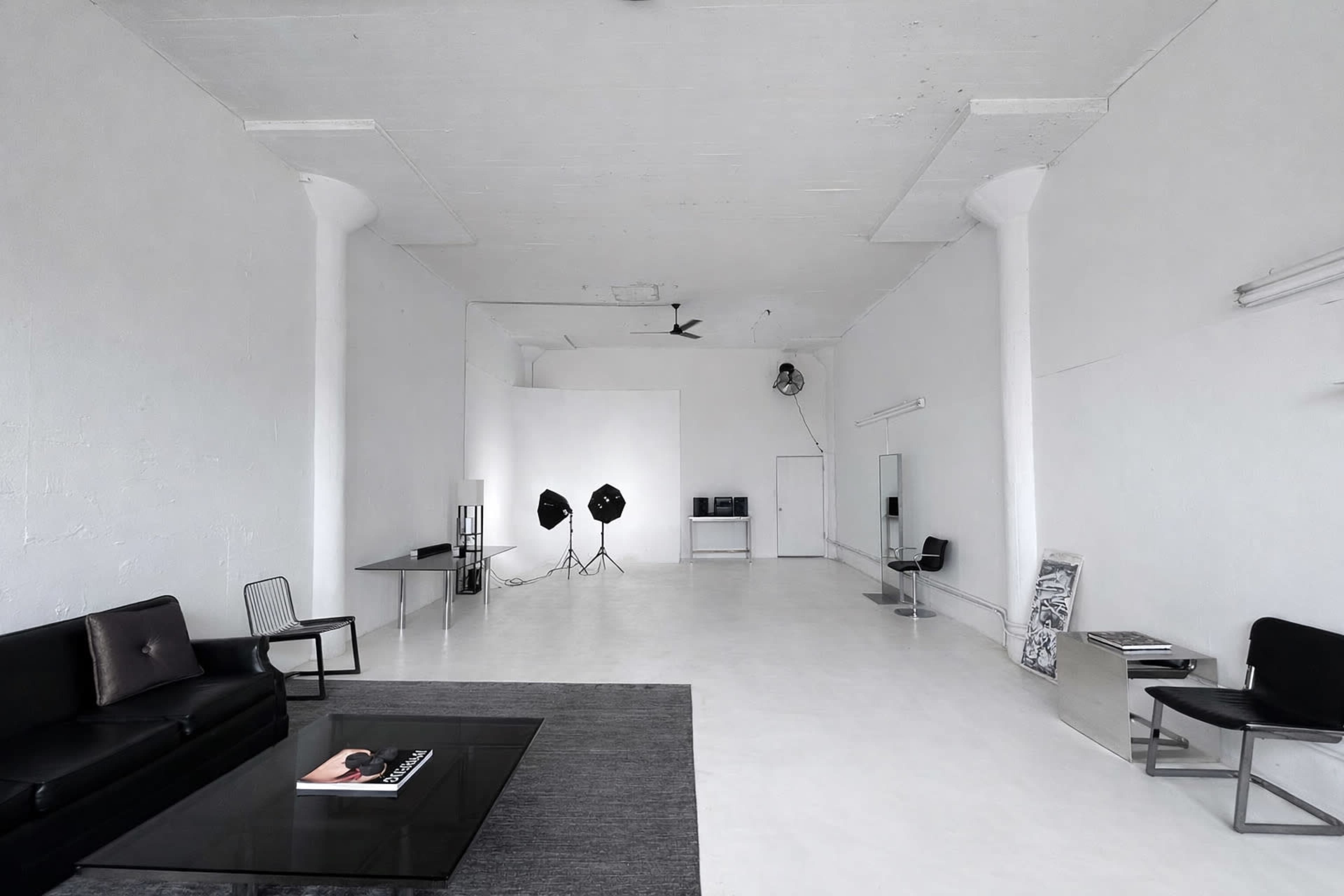 A spacious, minimally furnished room with white walls, a few chairs, a coffee table, and studio lighting equipment.