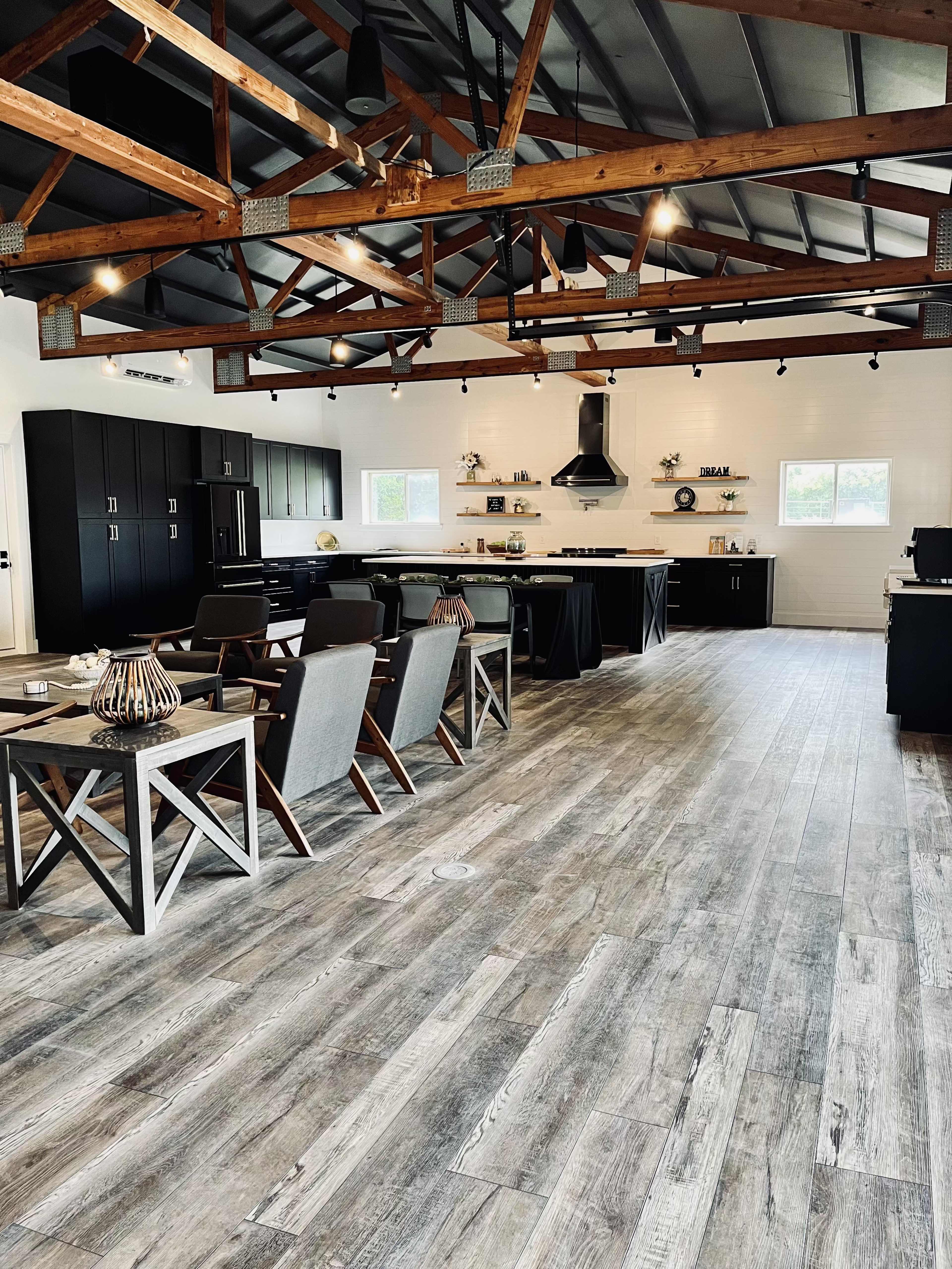 A modern kitchen and dining area with exposed wooden beams, black cabinetry, and a polished wooden floor, featuring seating arrangements and kitchen appliances.