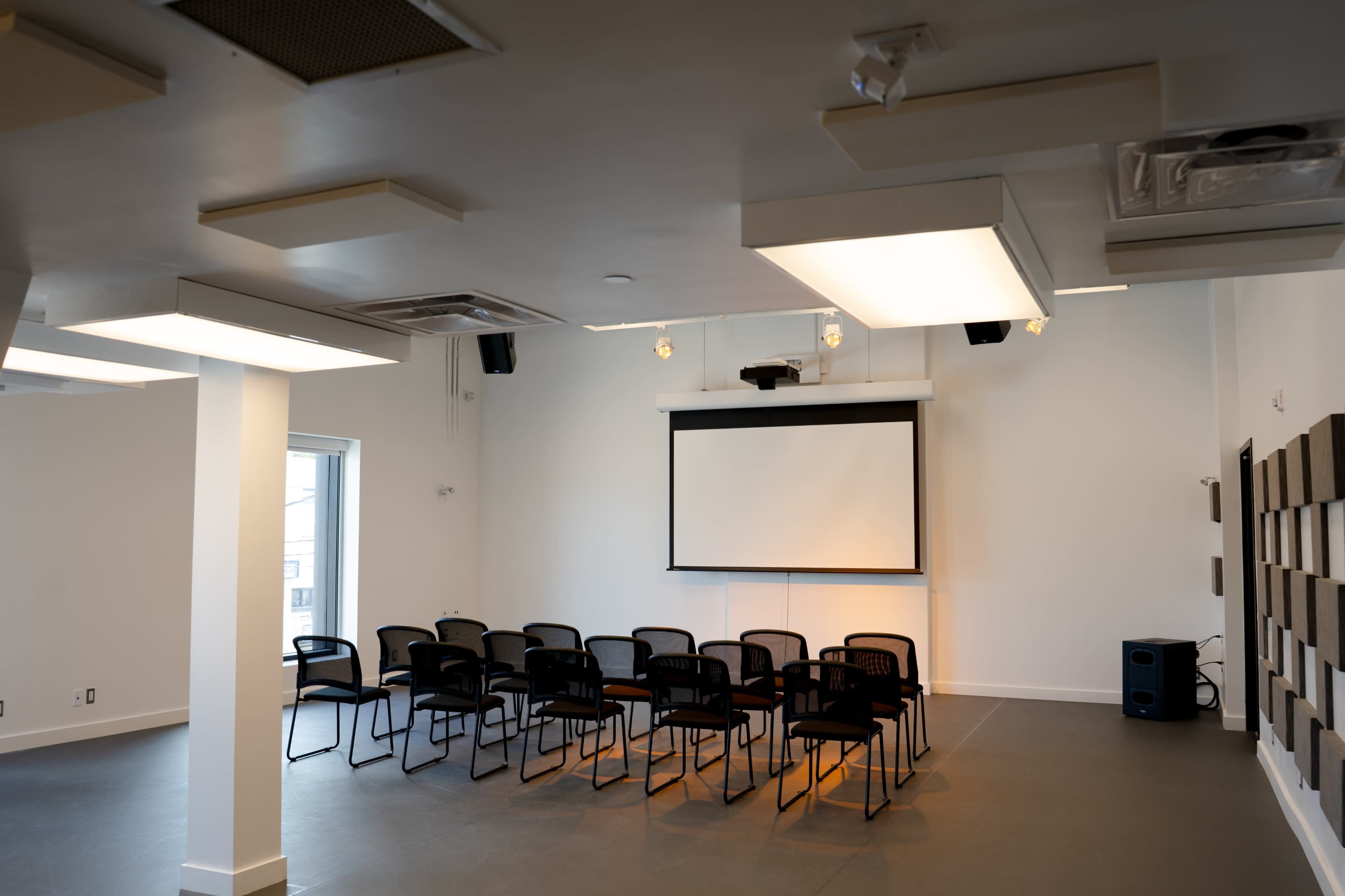 A meeting room features a row of black chairs arranged facing a large screen on the wall, with a projector mounted above.