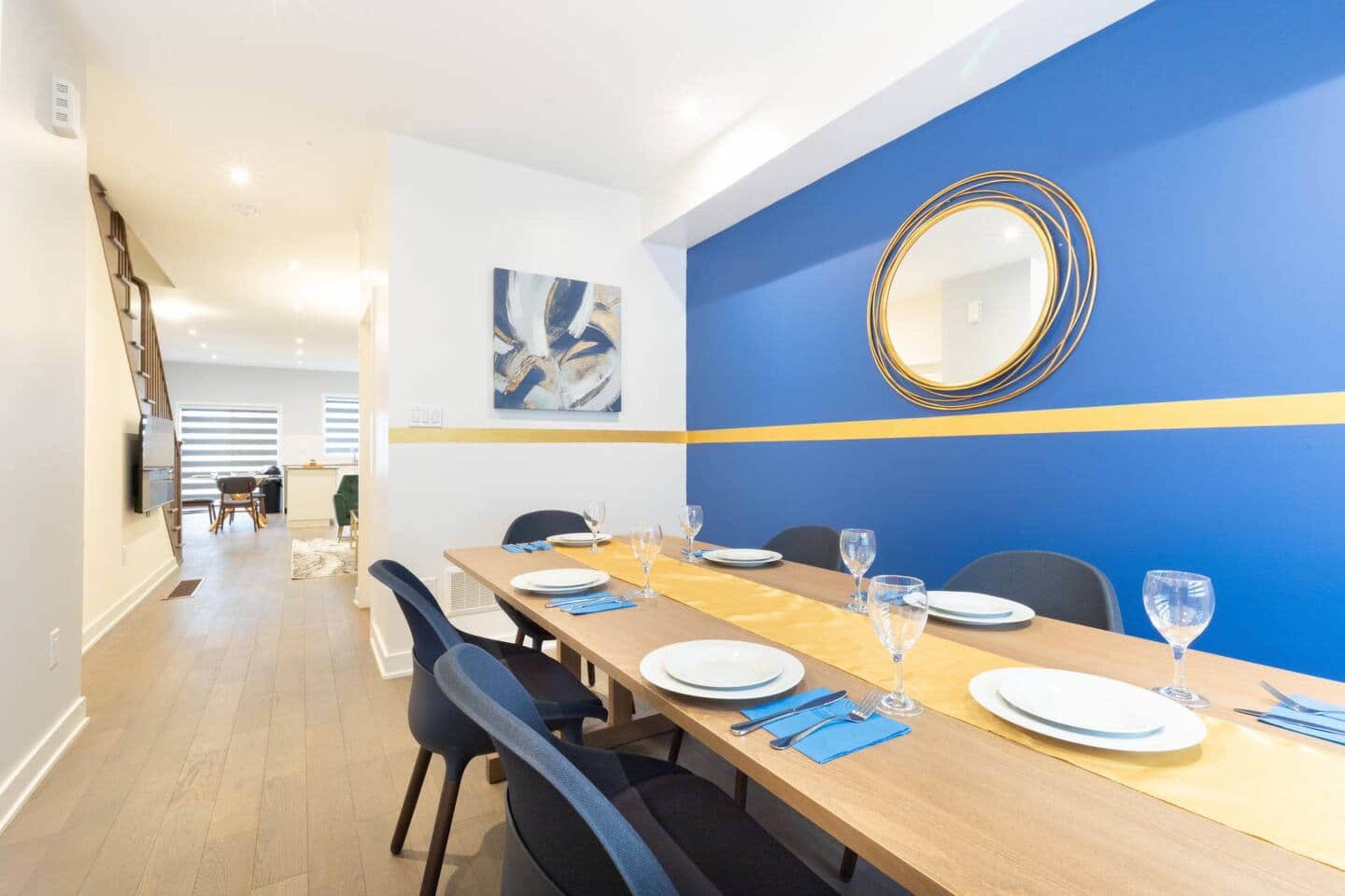 A dining area features a long wooden table set with plates and glasses, against a blue and white wall with a round mirror and artwork.