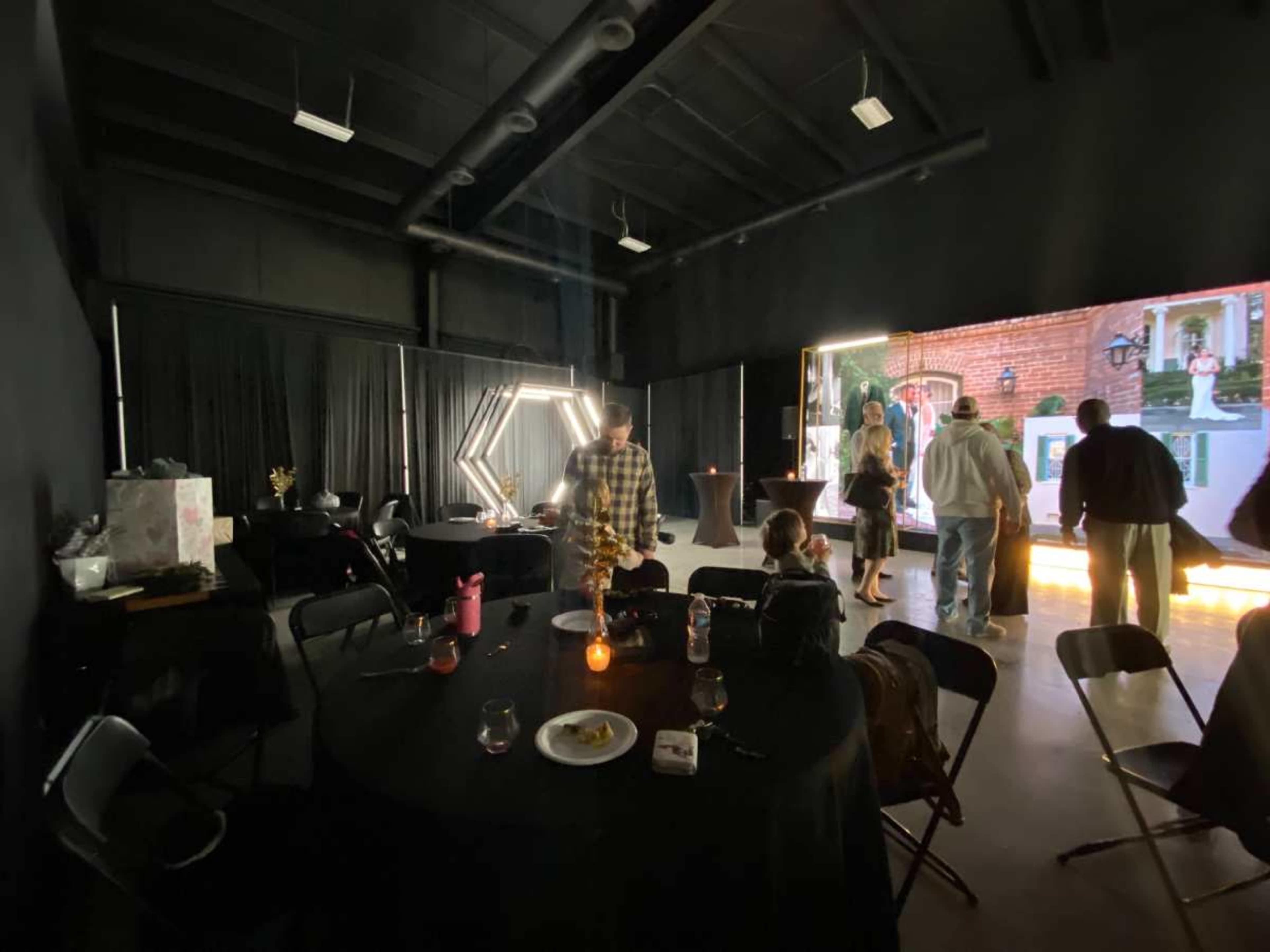 A dimly lit event space features tables with black linens and a large LED screen displaying imagery, while guests mingle throughout the room.